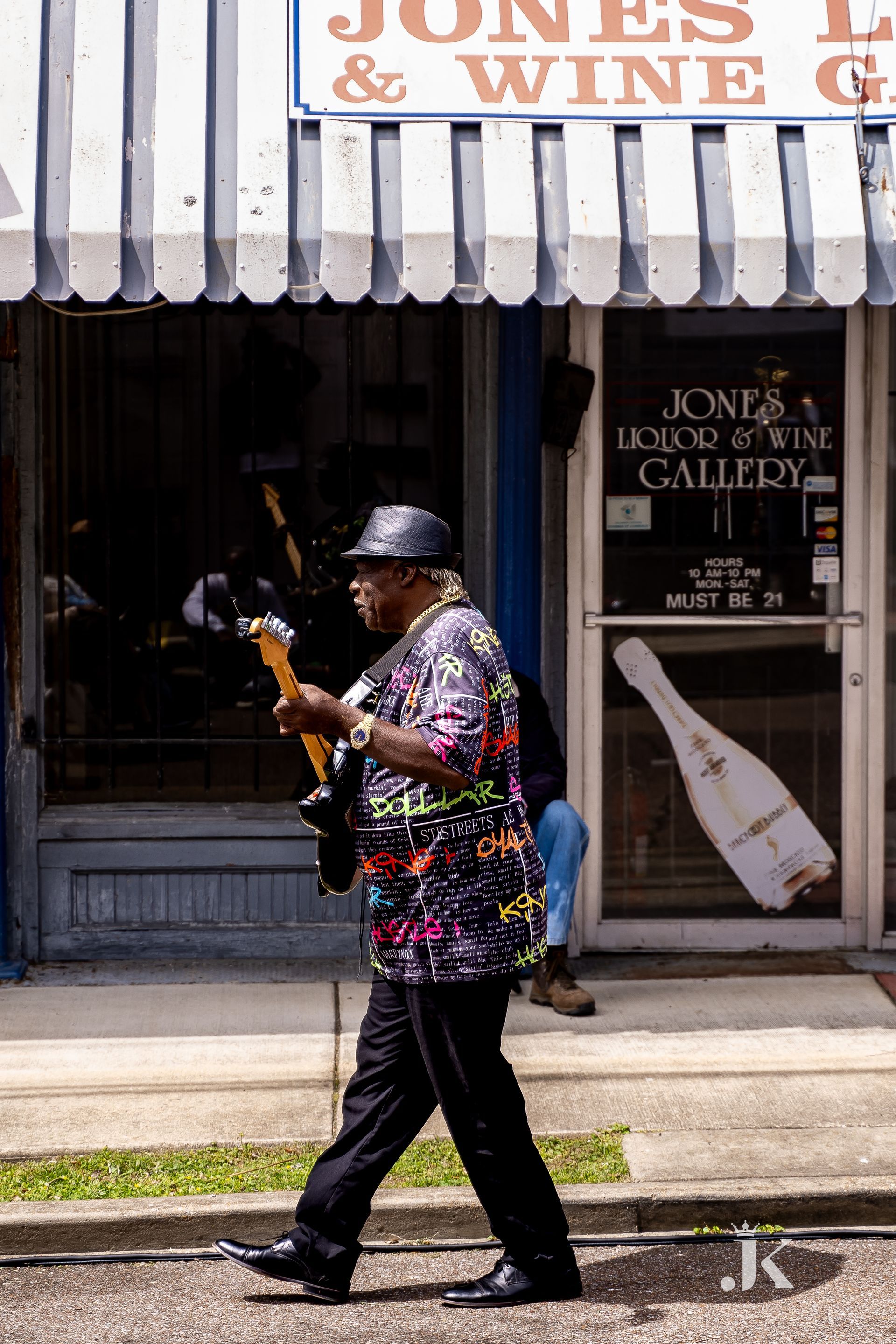 A man is walking down the street holding a guitar in front of a store called jones & wine.