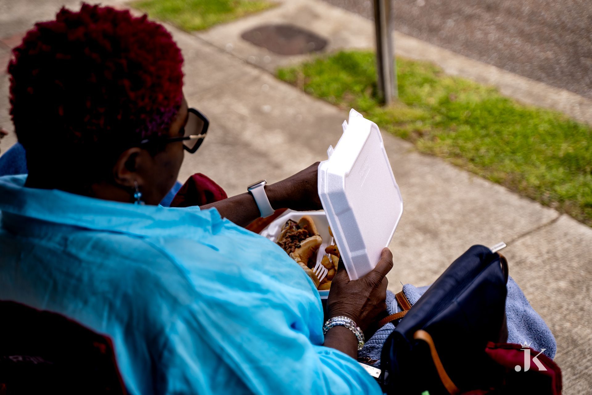 A woman is sitting on the sidewalk eating food from a styrofoam container.
