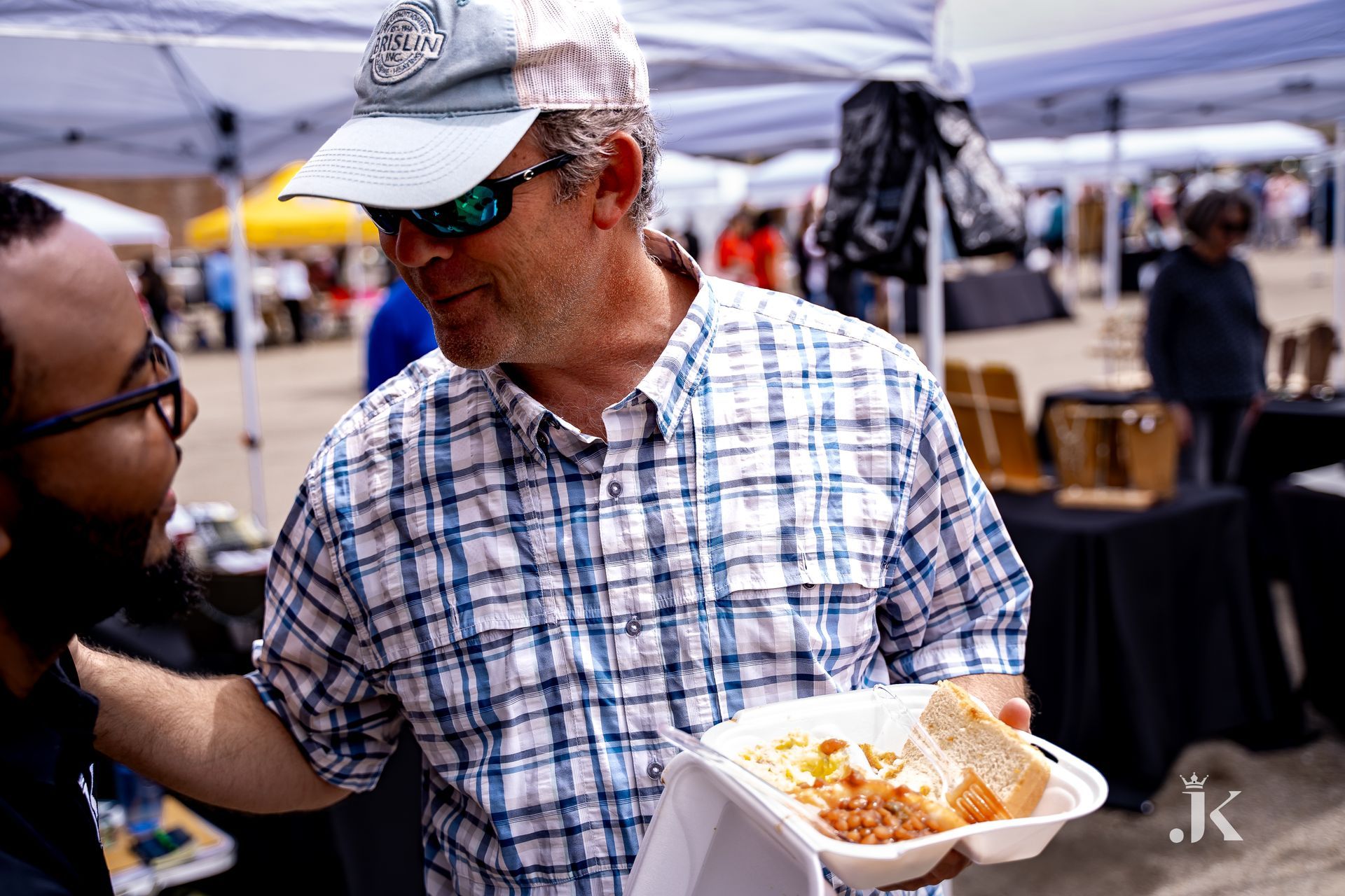 A man in a plaid shirt is holding a tray of food and talking to another man.