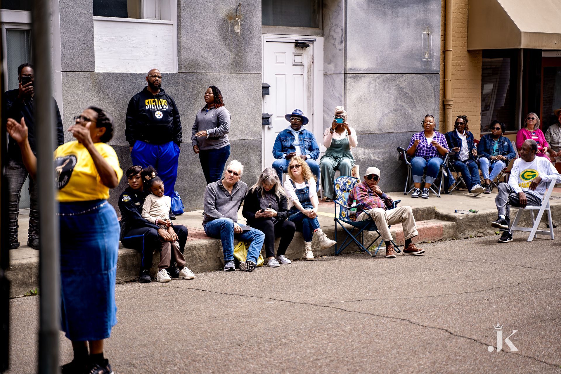 A group of people are sitting on the sidewalk watching a parade.