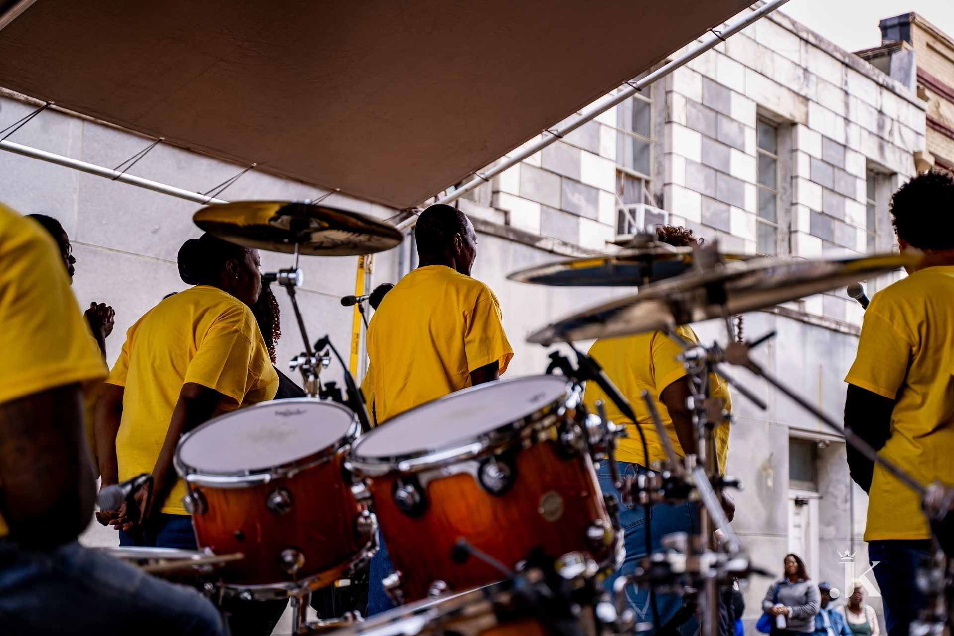 A group of people in yellow shirts are playing drums in front of a building.