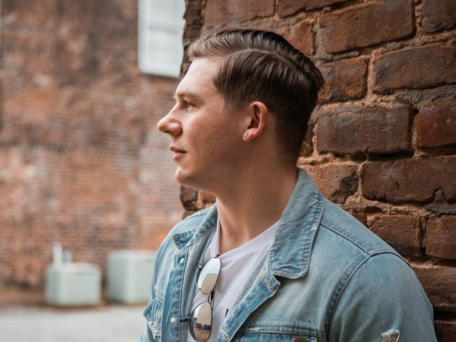 A man in a denim jacket is leaning against a brick wall.