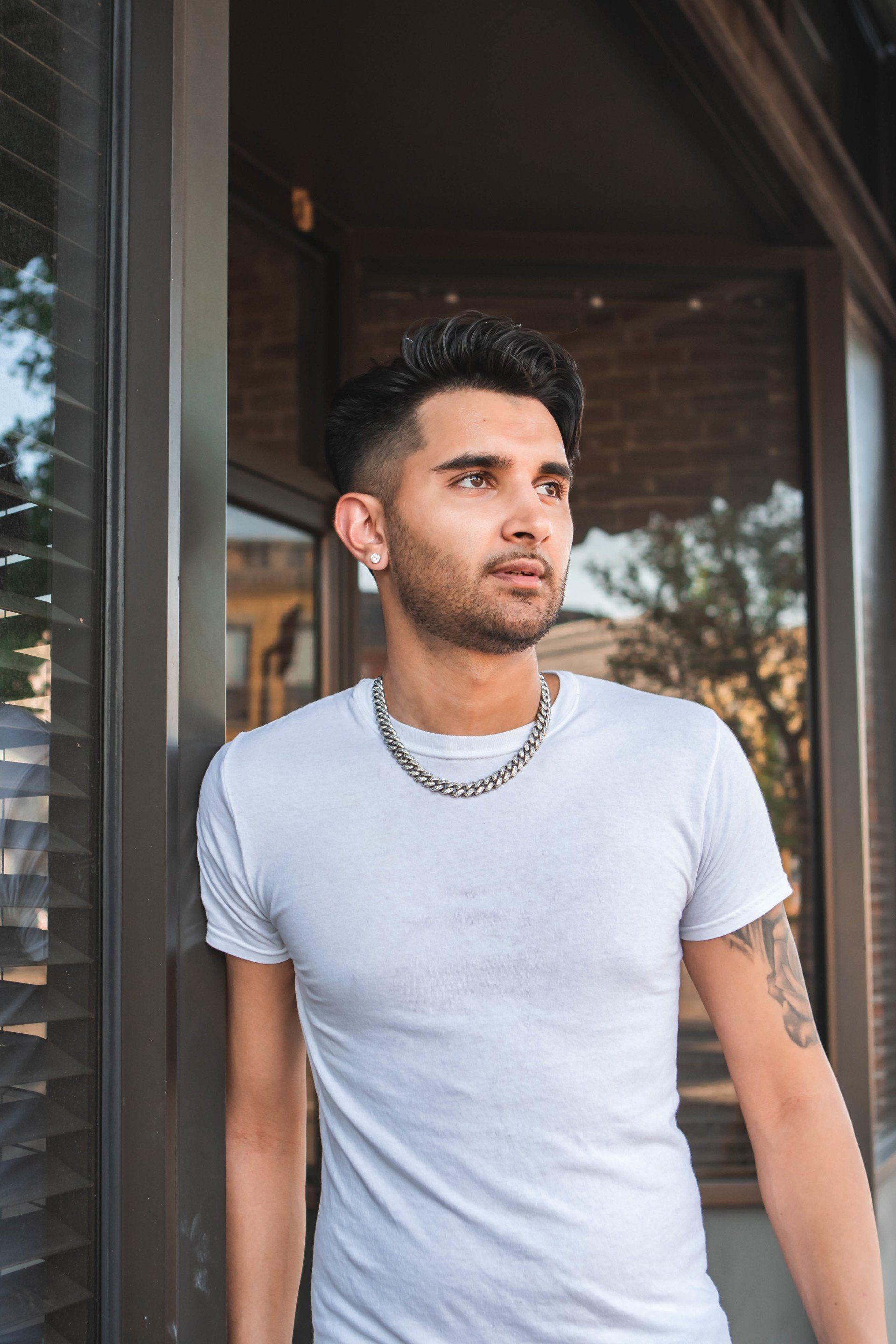 A man wearing a white t-shirt and a chain necklace is standing in front of a window.