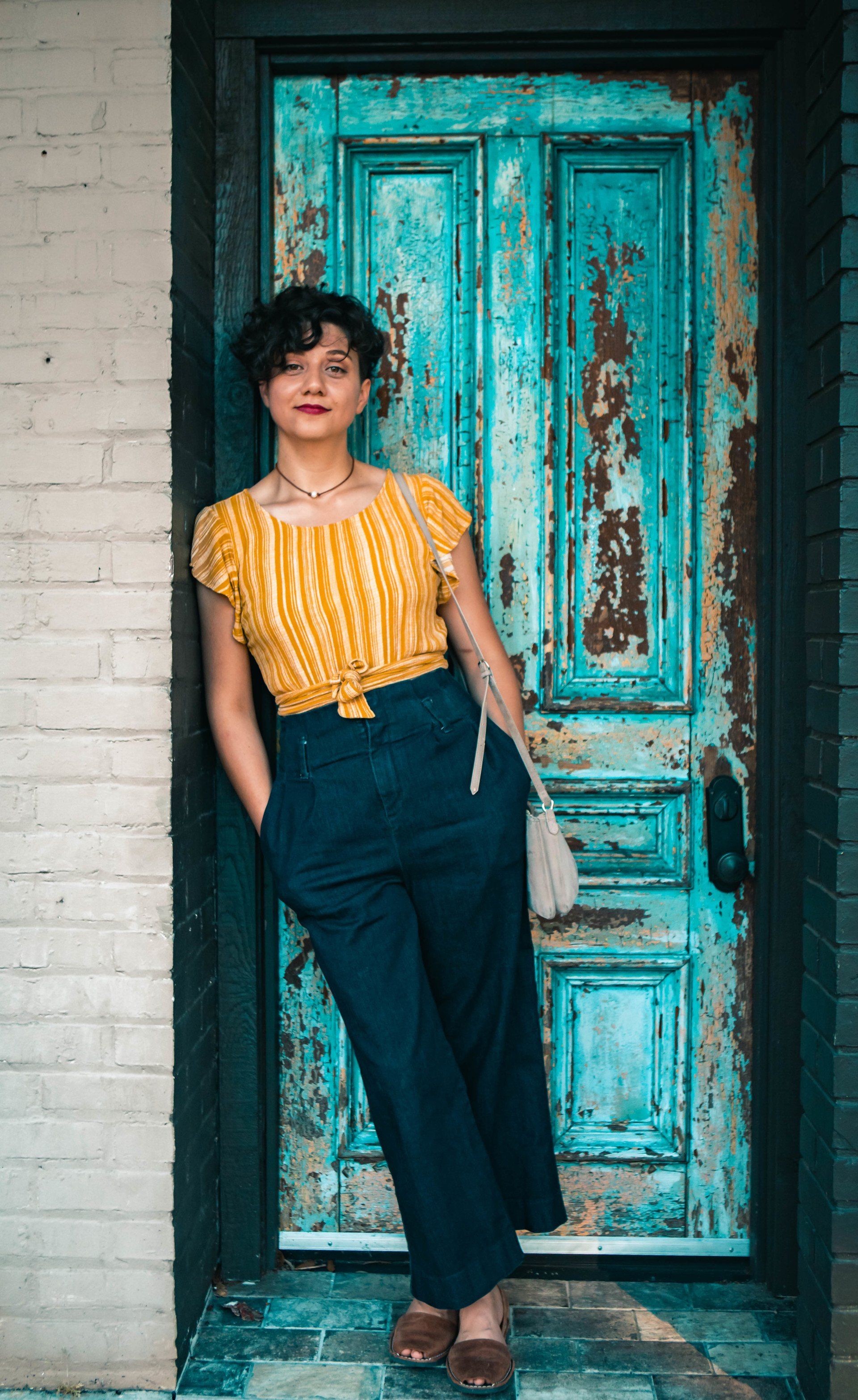 A woman in a yellow top and blue pants is standing in front of a blue door.