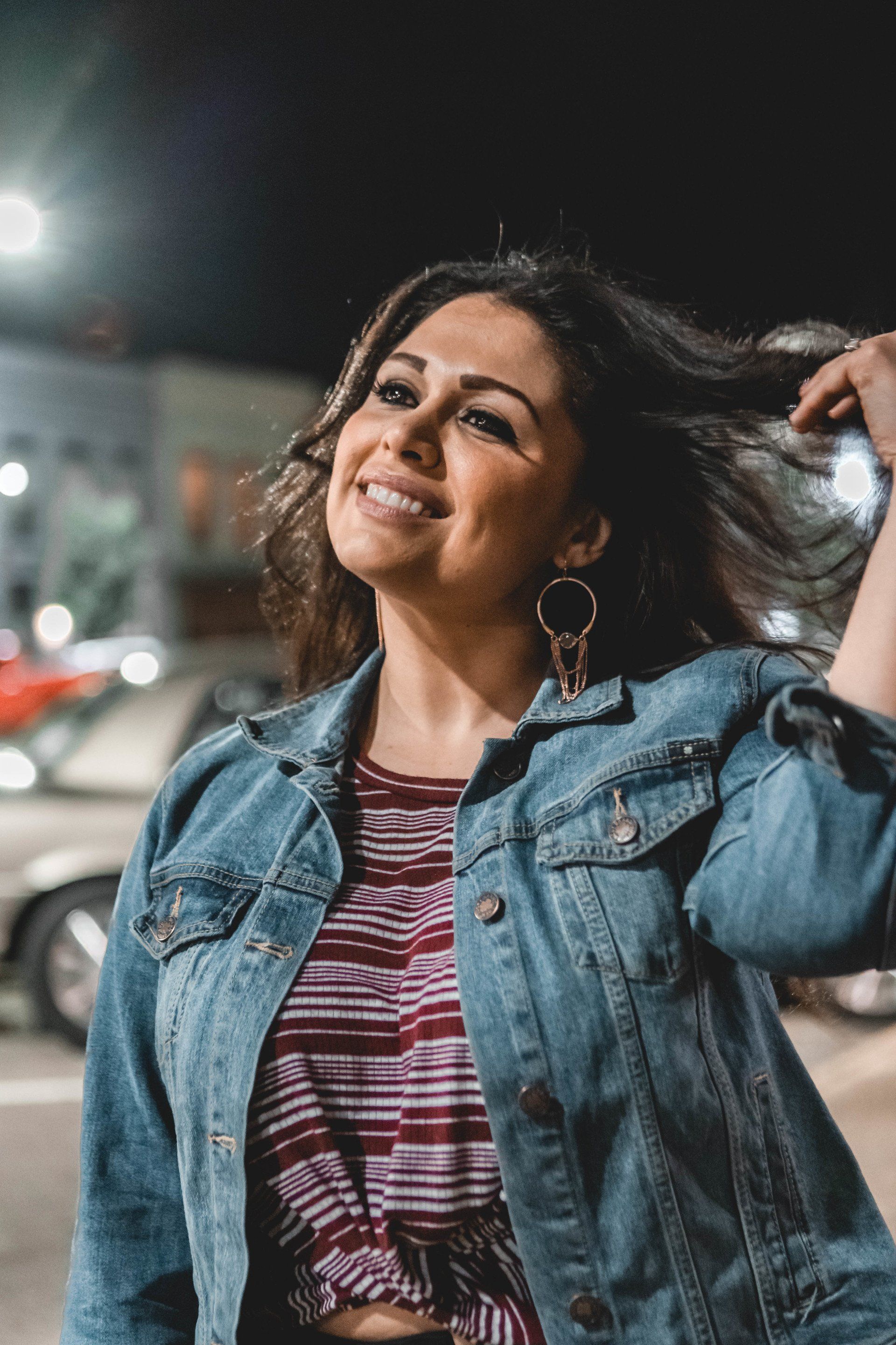 A woman in a denim jacket is smiling and holding her hair.