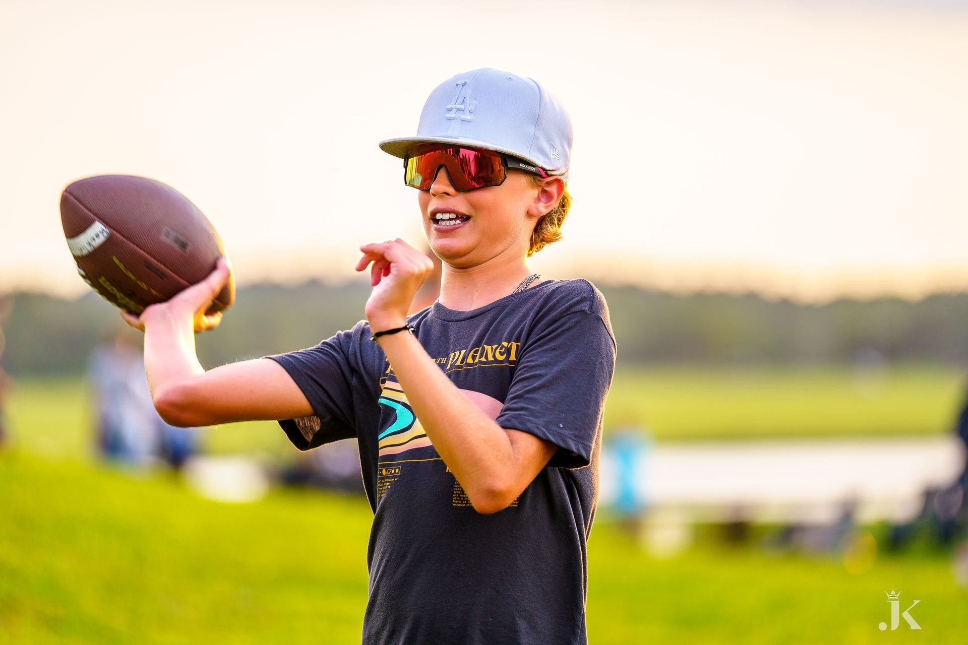 A young boy wearing sunglasses and a baseball cap is throwing a football.