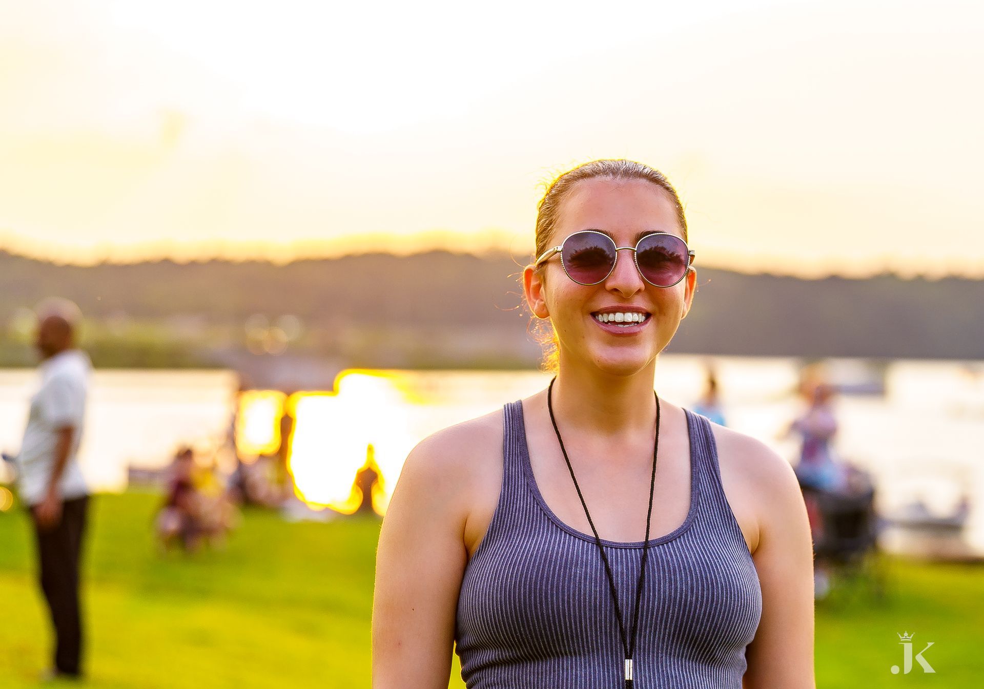 A woman wearing sunglasses and a tank top is standing in front of a lake.