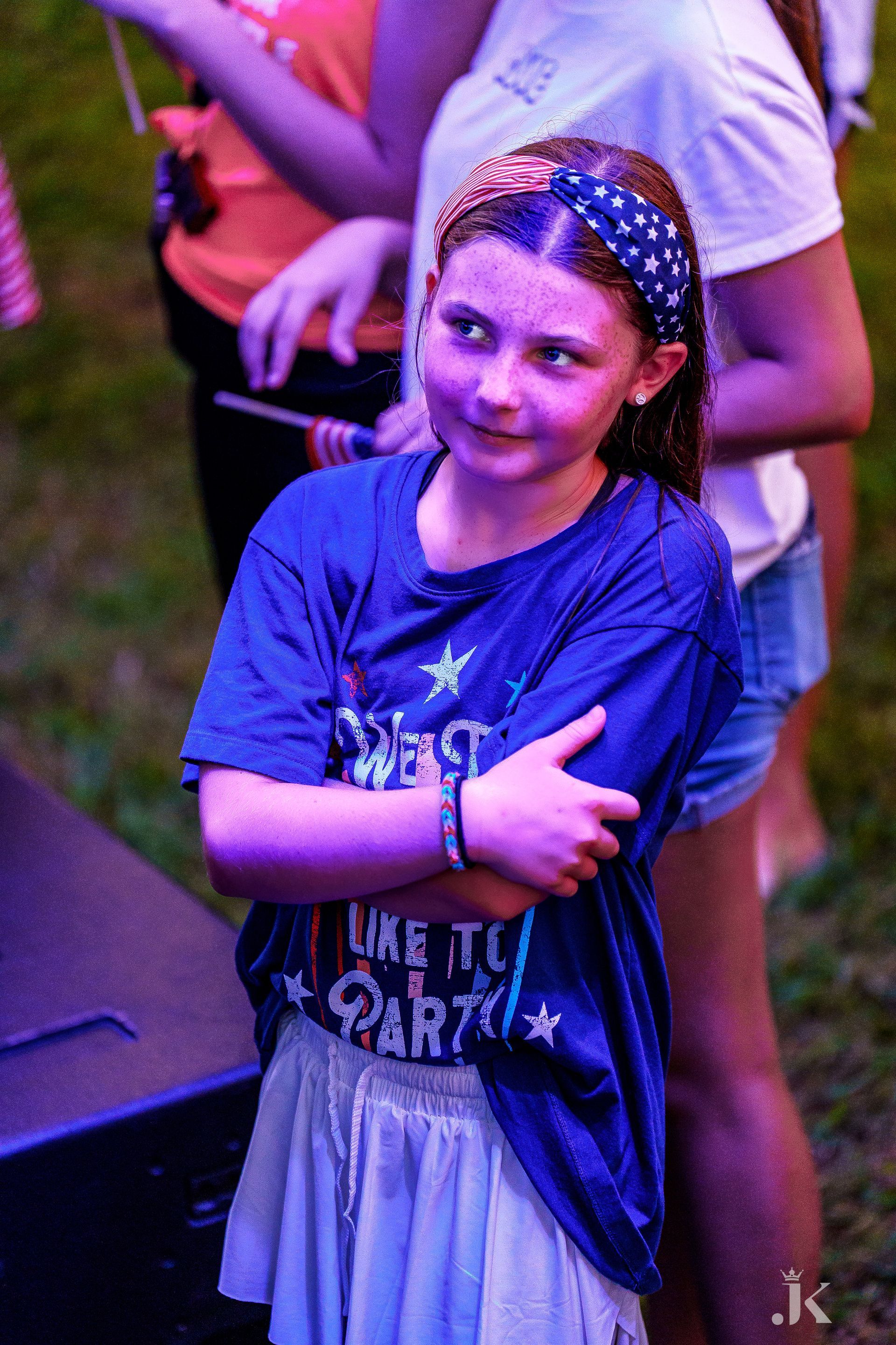 A young girl wearing a blue shirt and a headband is standing in front of a stage.