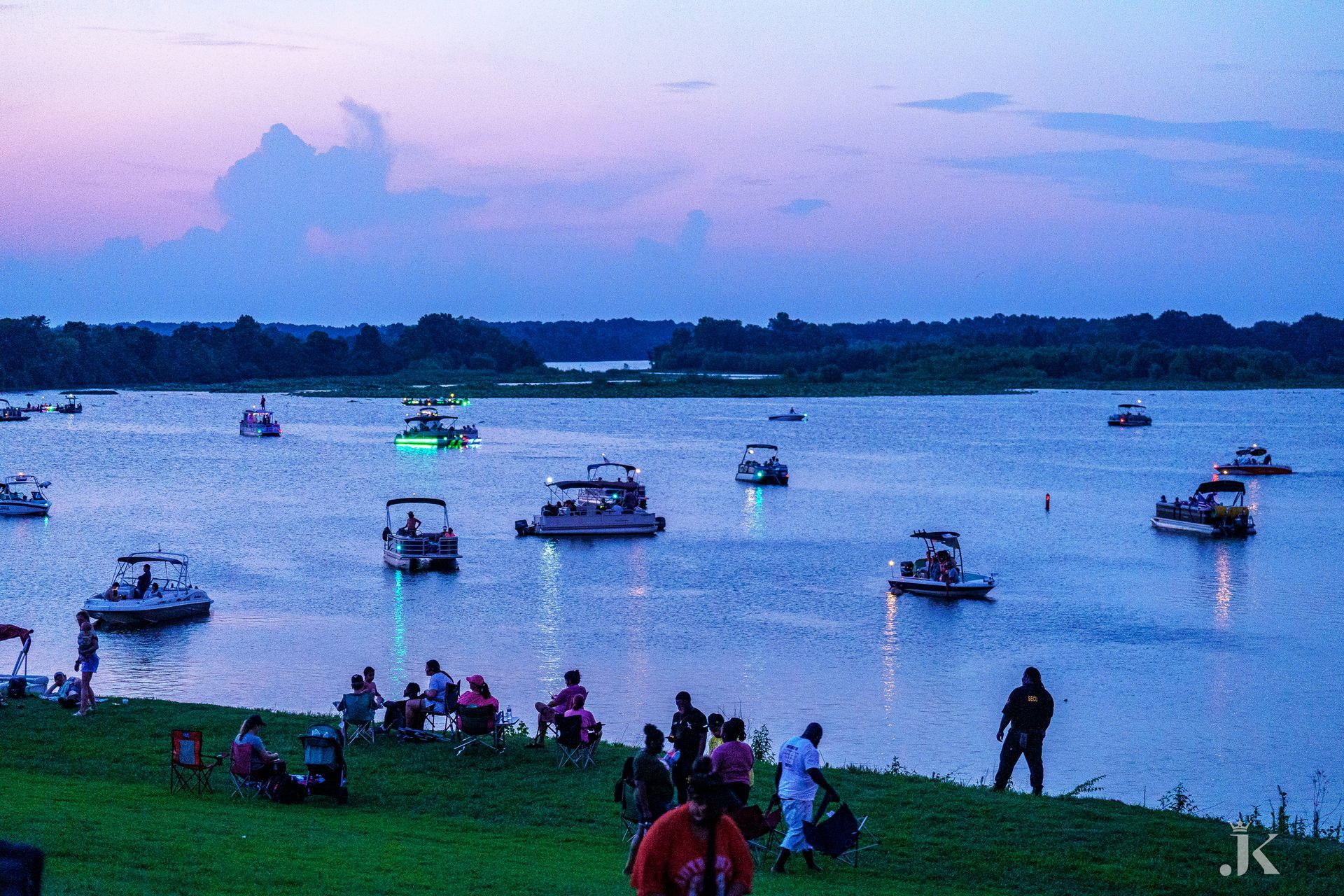 A group of people are sitting on the shore of a lake with boats in the water.