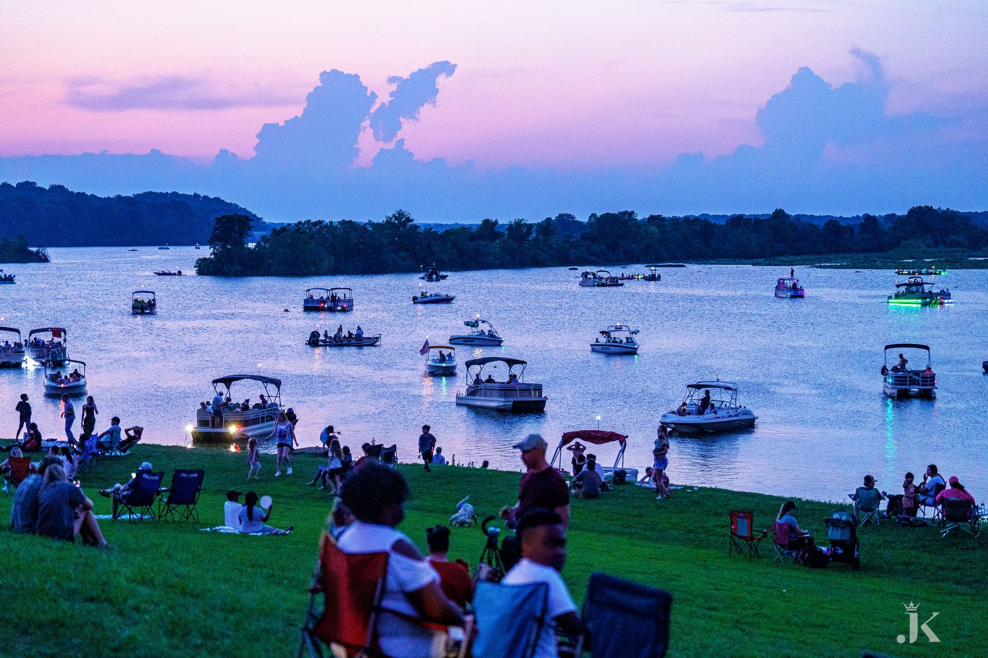 A group of people are sitting on the shore of a lake with boats in the water.