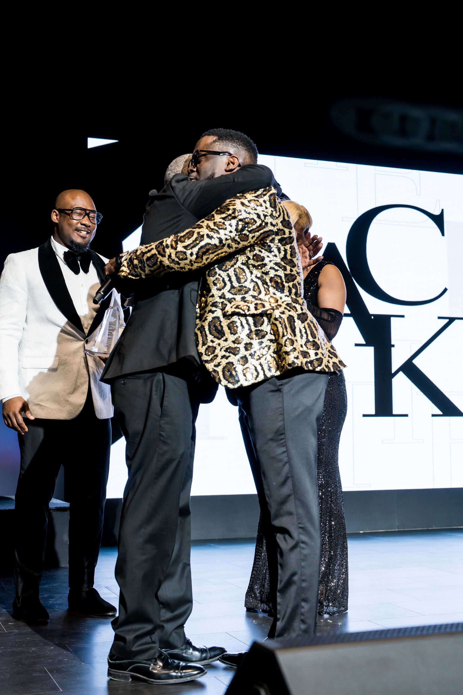 A man in a leopard print jacket is hugging another man on a stage.