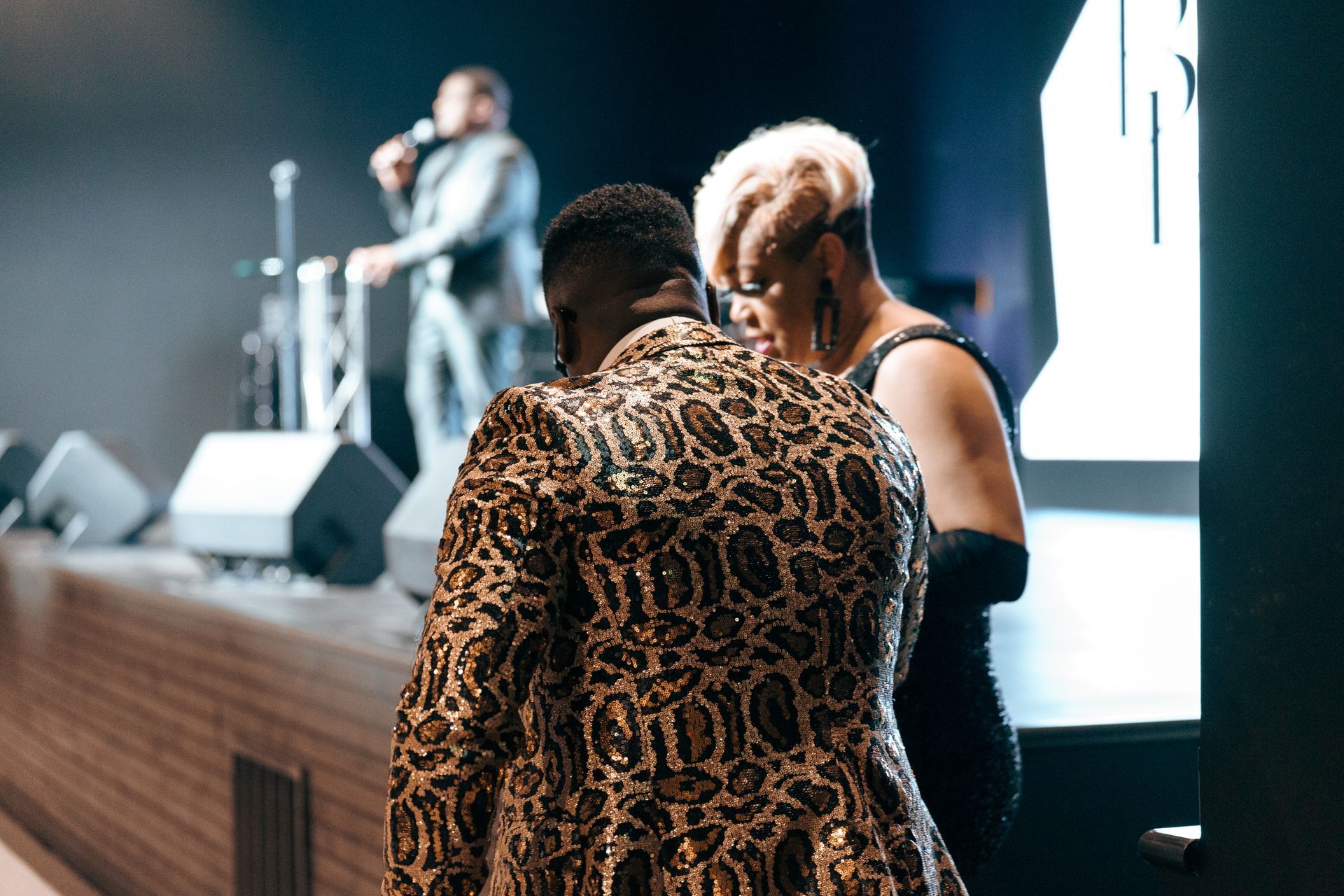 A man in a leopard print jacket is kneeling down next to a woman on a stage.