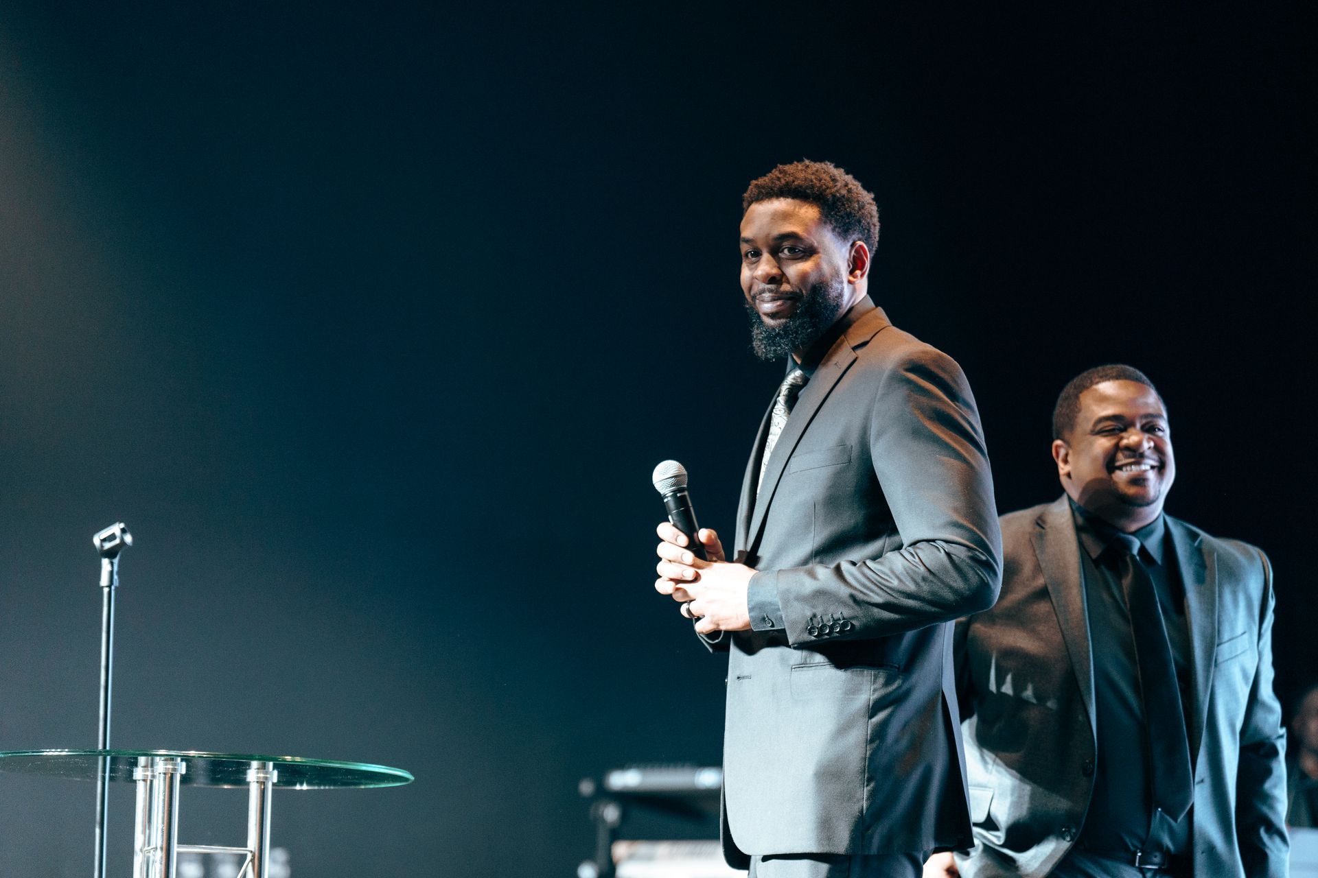 Two men in suits are standing on a stage holding microphones.