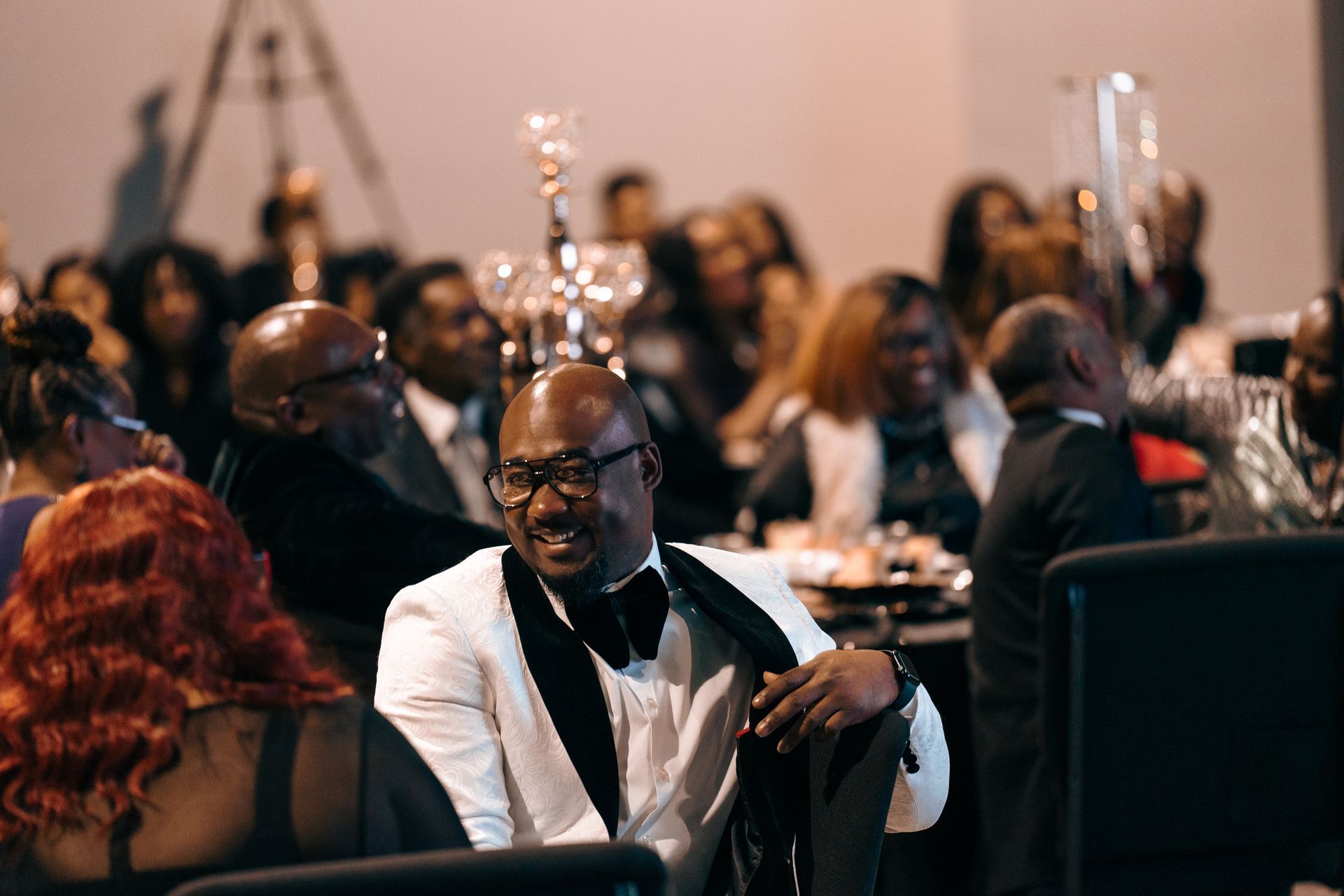 A man in a tuxedo is sitting in front of a crowd of people at a dinner party.
