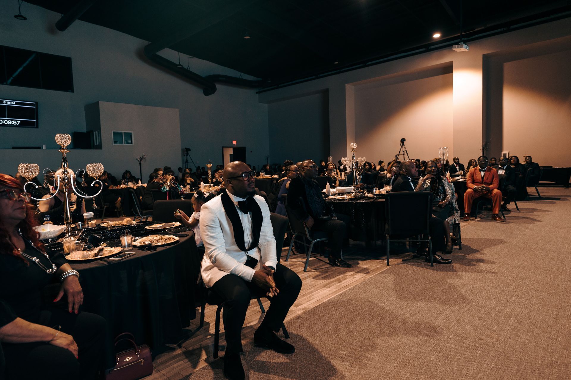 A man in a tuxedo is sitting in a chair in a large room.