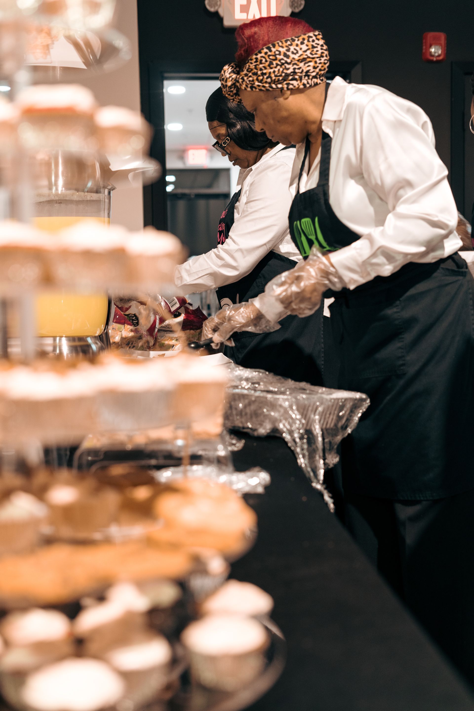 Two women are preparing food at a table with cupcakes.