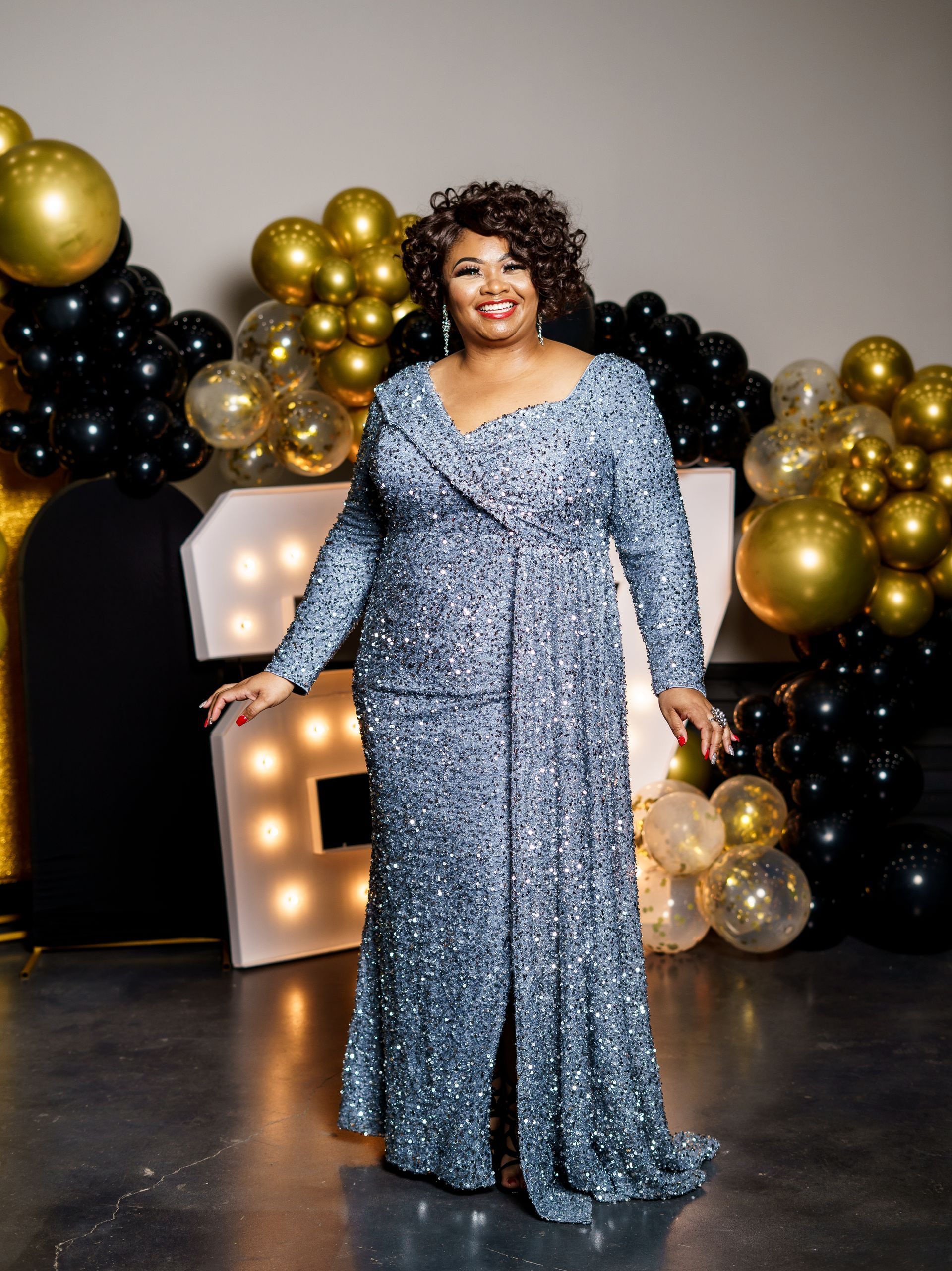A woman in a long blue sequined dress is standing in front of balloons.