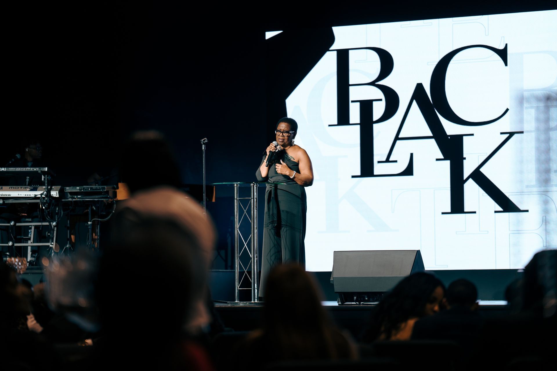 A woman is standing on a stage with a microphone in front of a large screen that says pac lk.