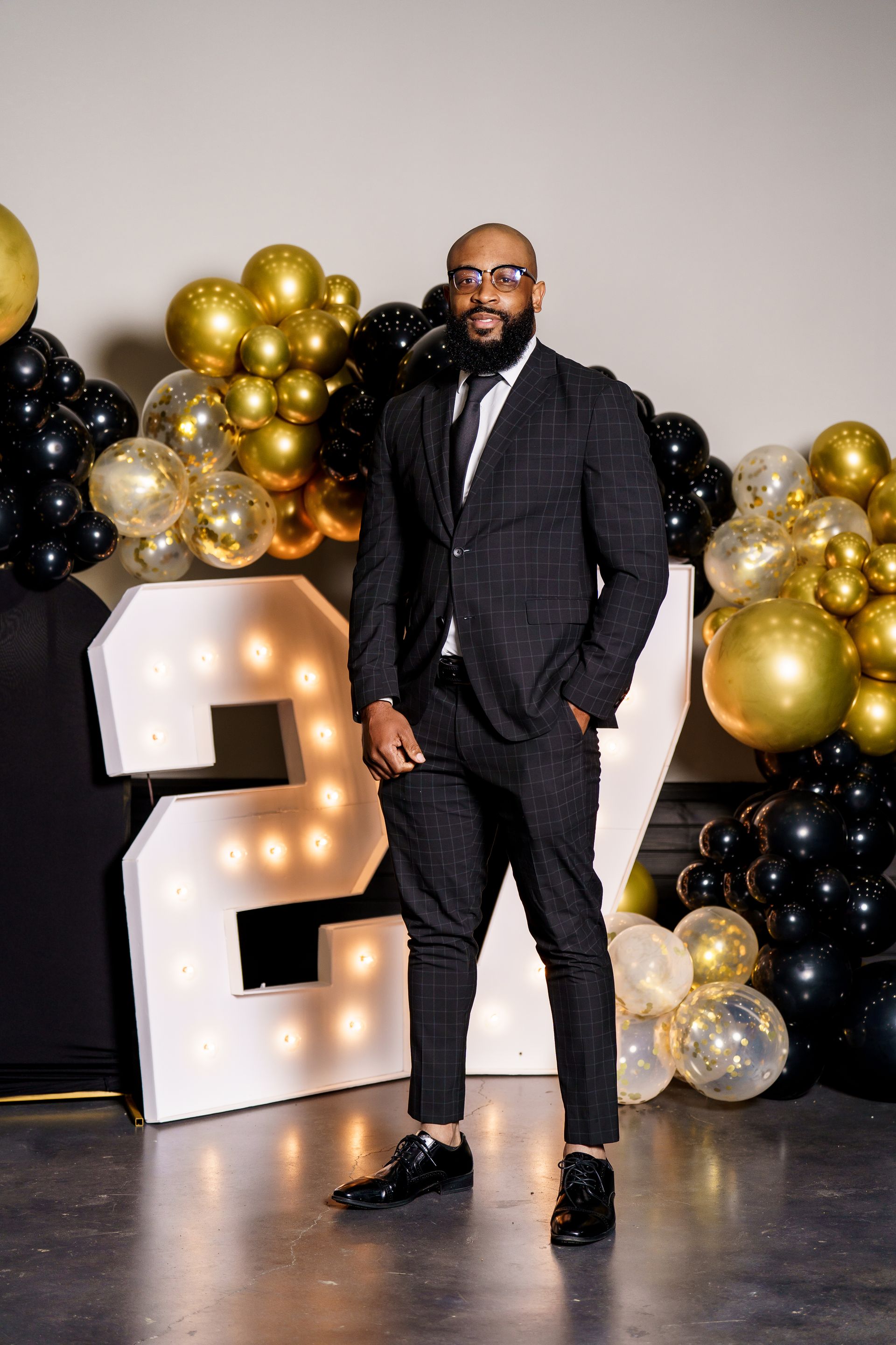 A man in a suit is standing in front of a large number 2 surrounded by balloons.