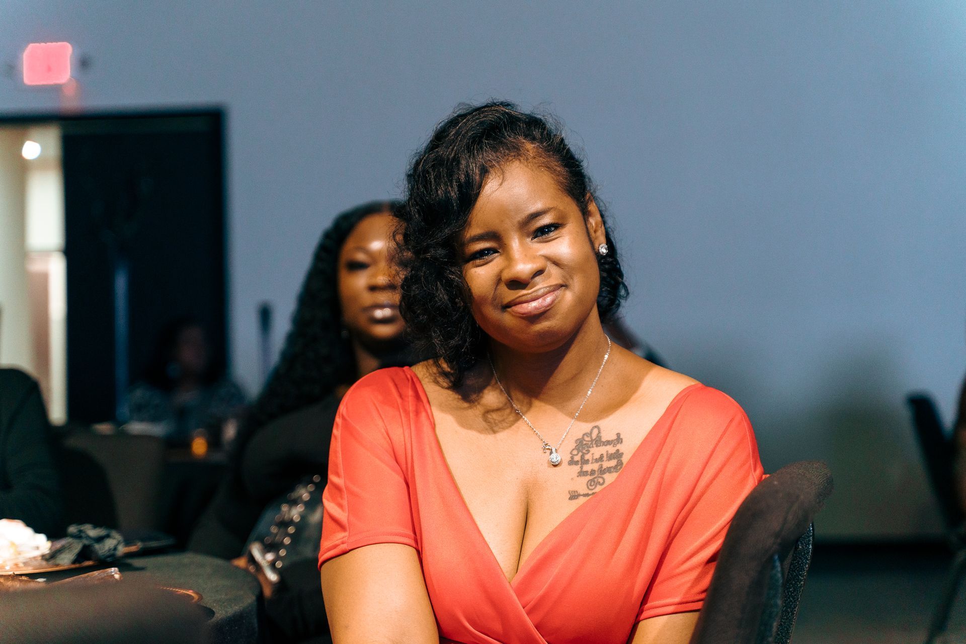 A woman in a red dress with a tattoo on her chest is sitting in a chair.