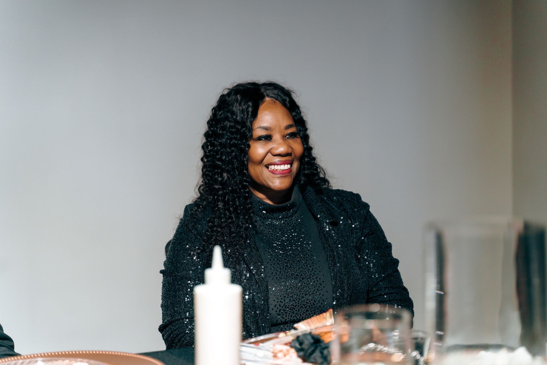 A woman in a black dress is sitting at a table with a bottle of ketchup.