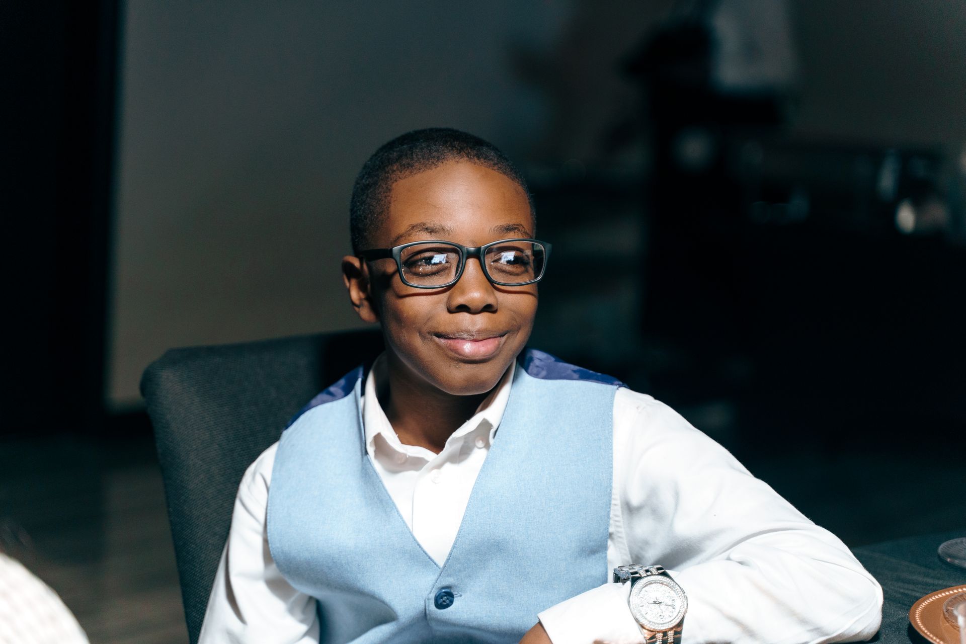 A young boy wearing glasses and a blue vest is sitting at a table.
