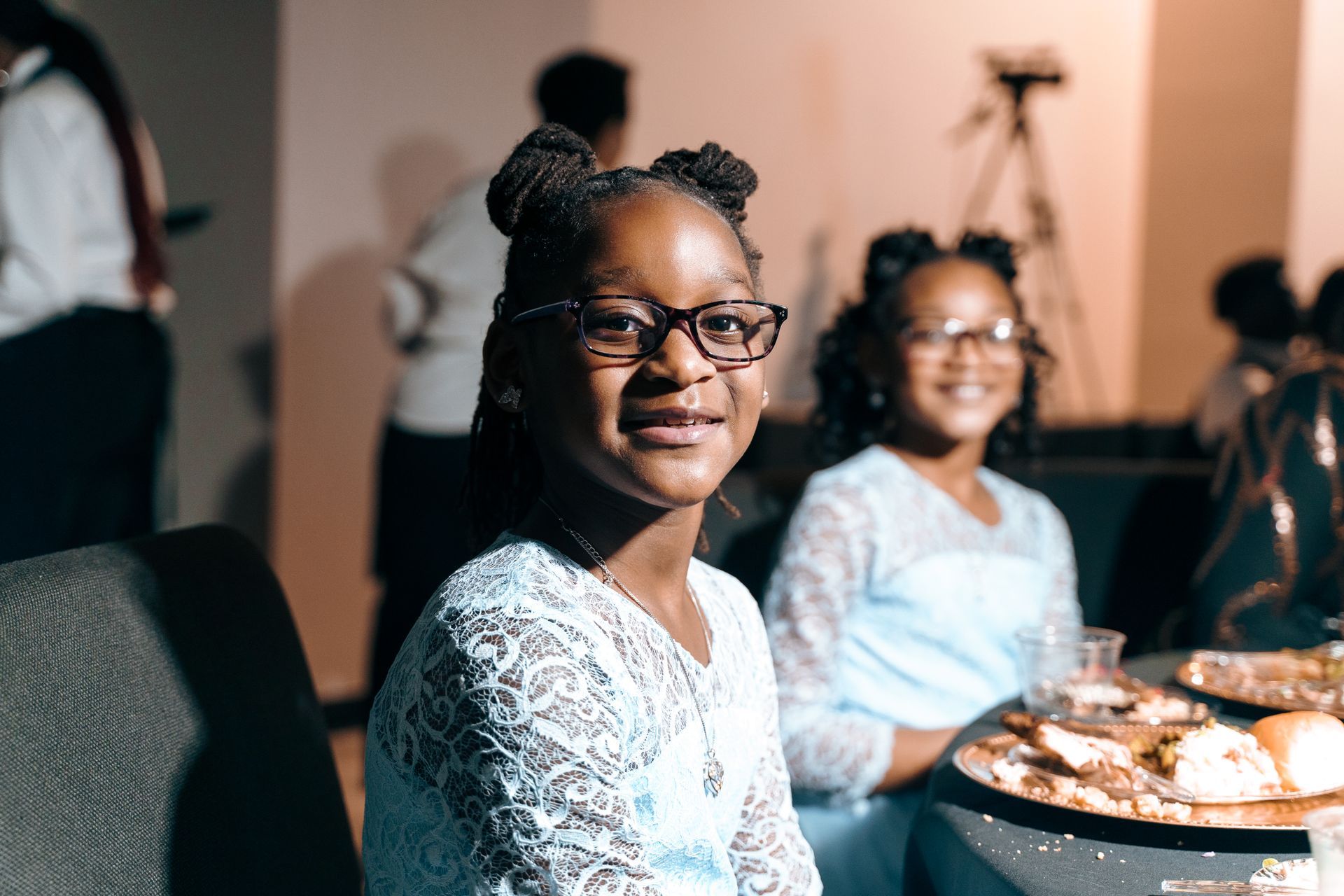 A little girl wearing glasses is sitting at a table with a plate of food.