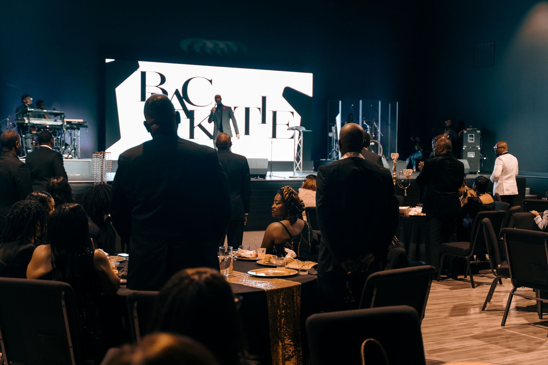 A group of people are sitting at tables in front of a large screen.