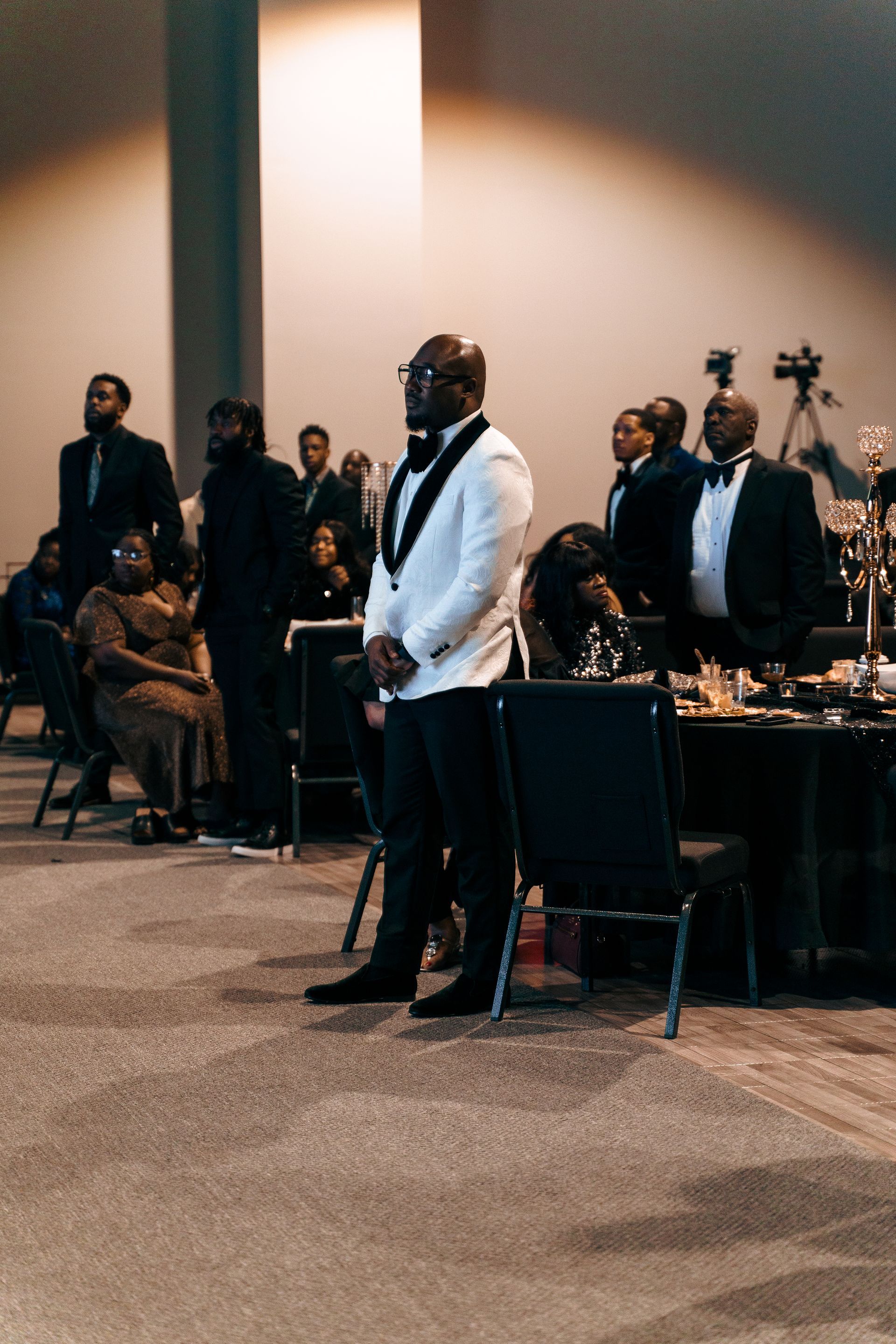 A man in a tuxedo is standing in a room with people sitting at tables.