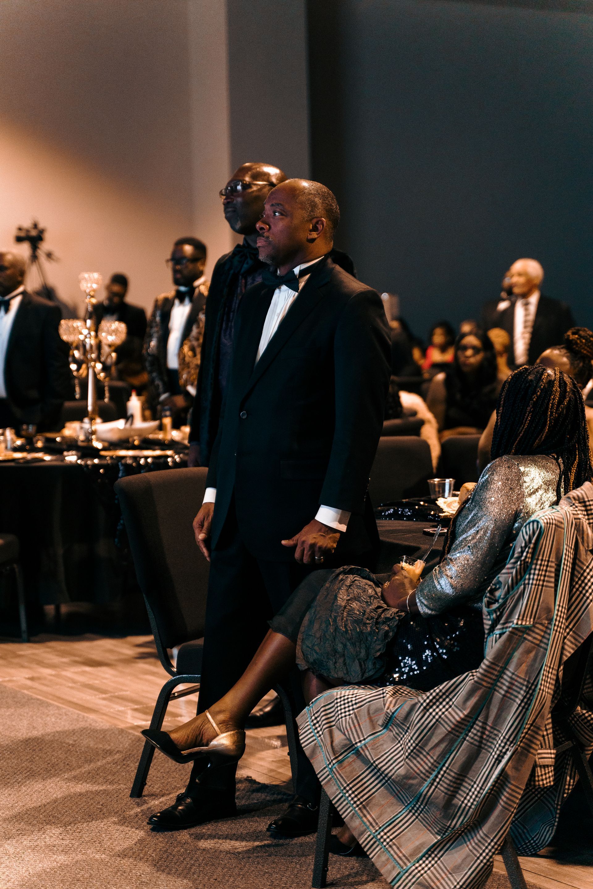 A man in a tuxedo is standing next to a woman in a chair.