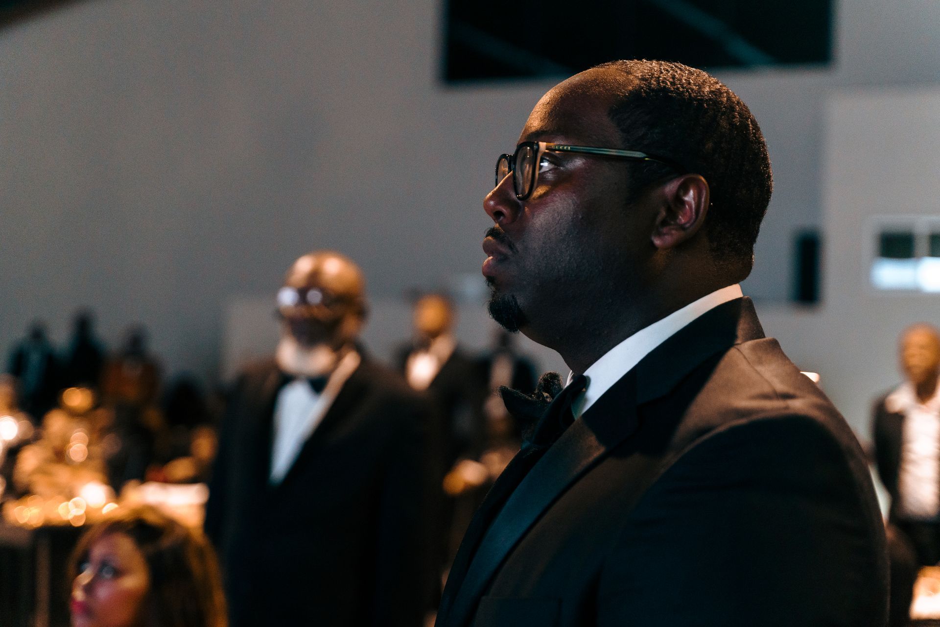 A man in a tuxedo and glasses is standing in front of a crowd.