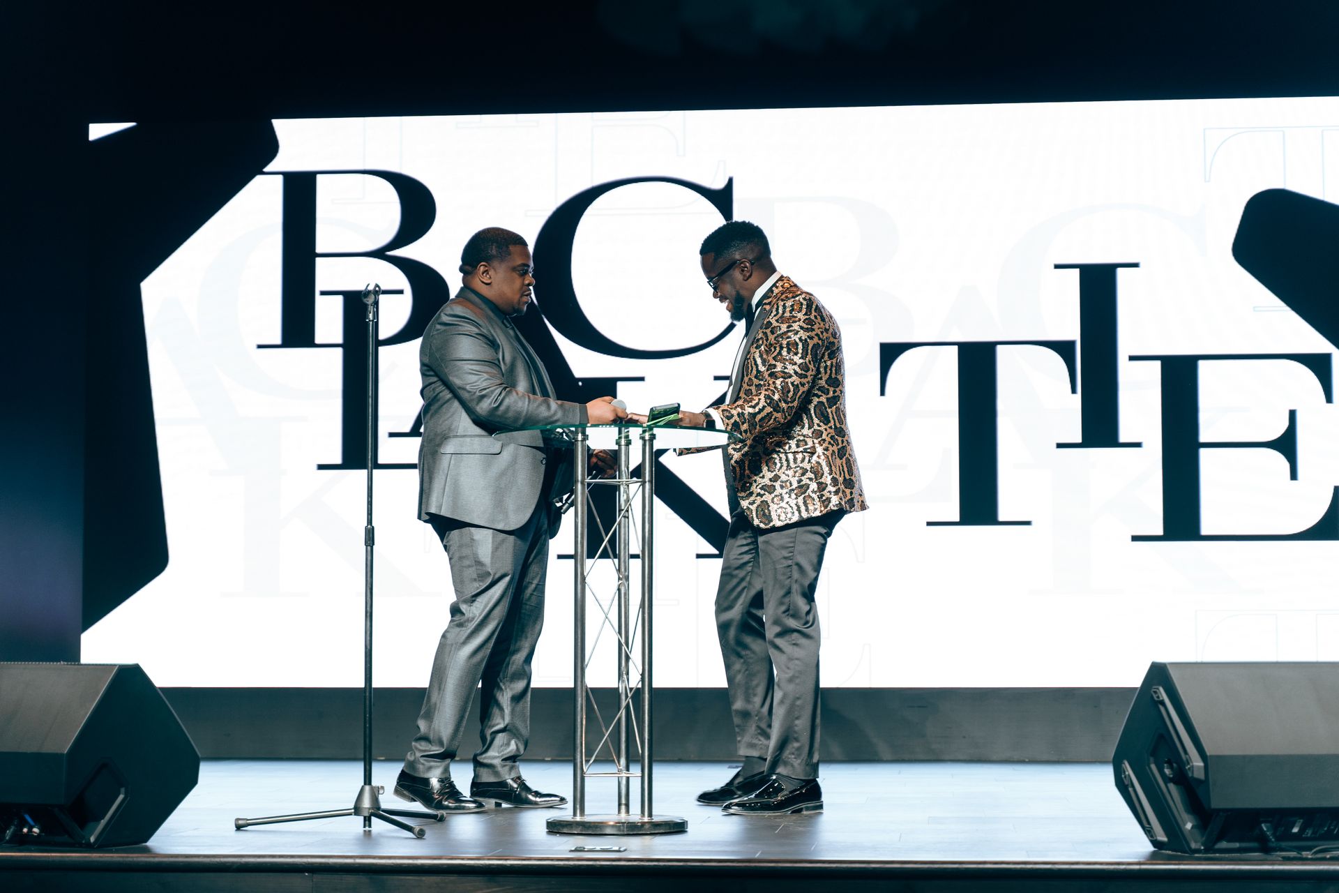 Two men are standing on a stage in front of a large screen.