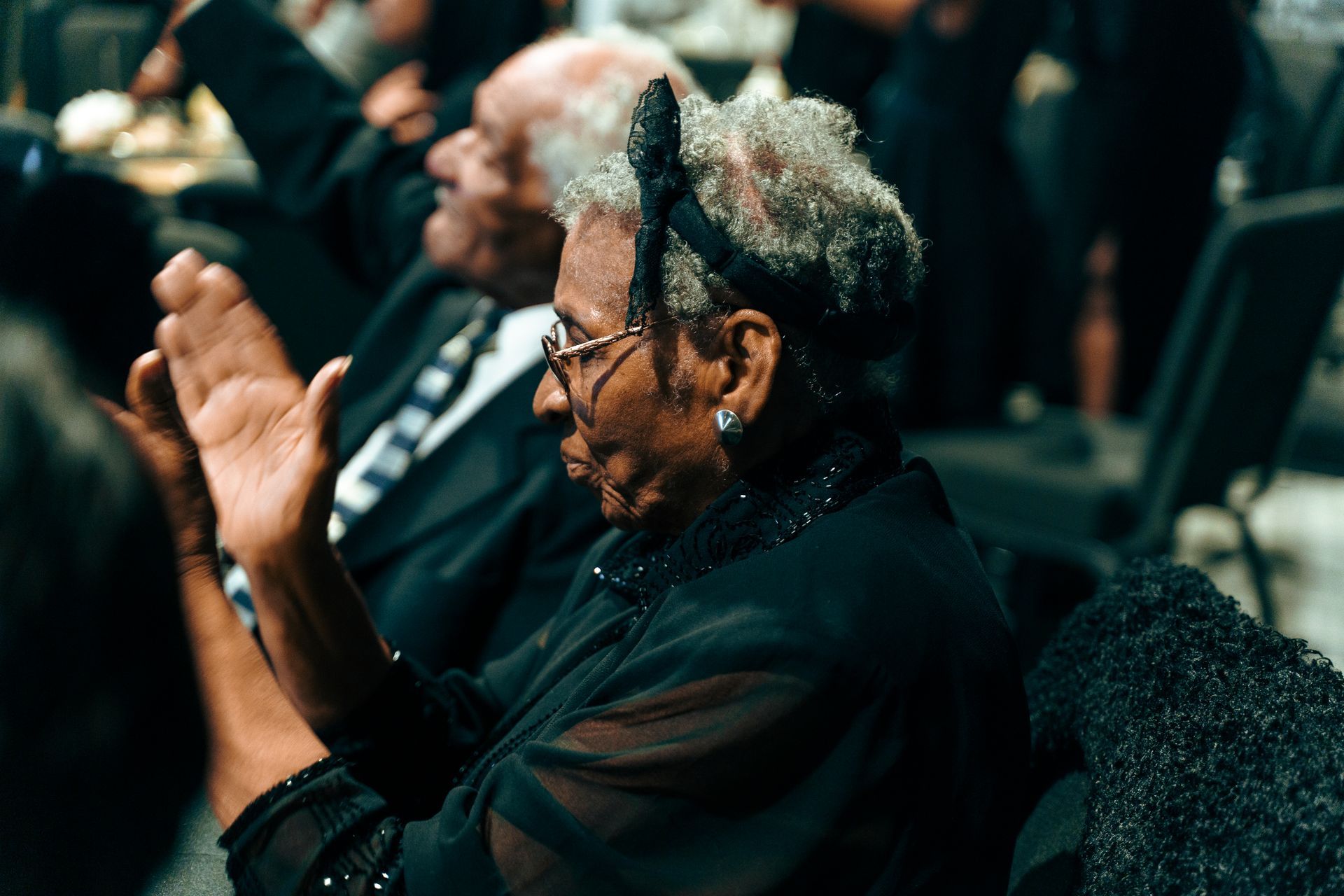 A woman in a black dress is sitting in a crowd clapping her hands.