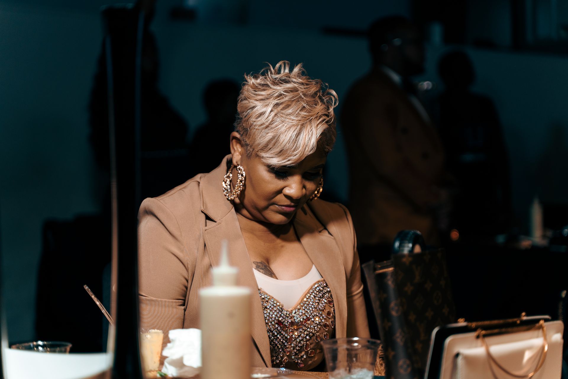 A woman is sitting at a table with a glass of water and a bottle of sauce.