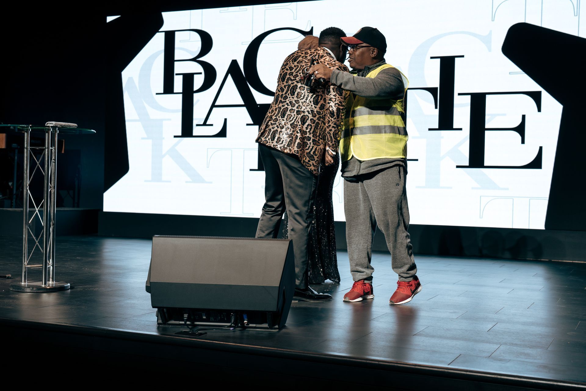 A man in a yellow vest is hugging another man on a stage.
