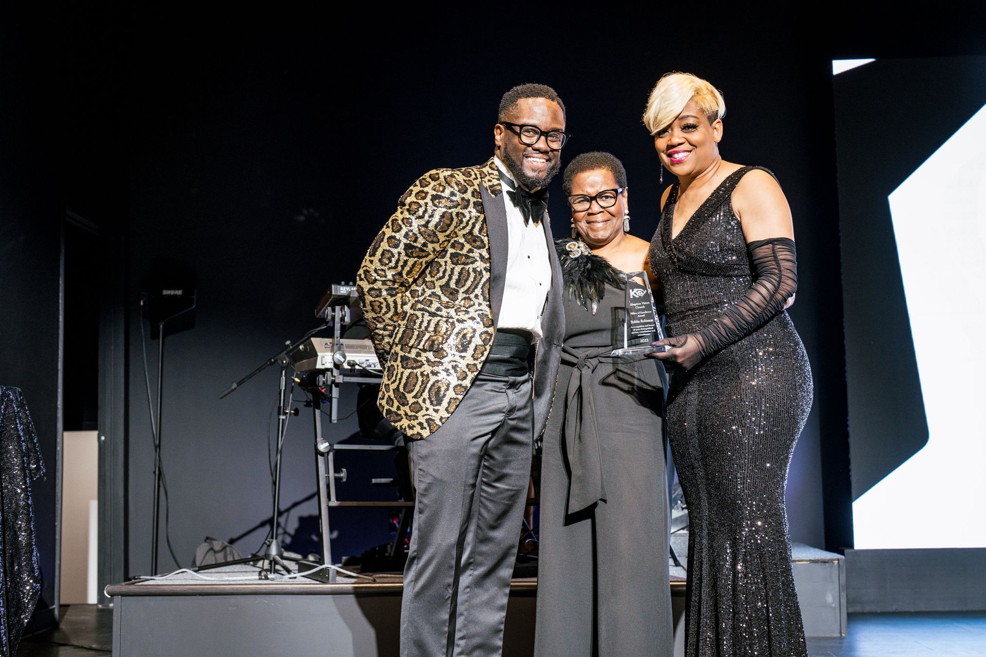 A man and two women are standing on a stage holding a trophy.