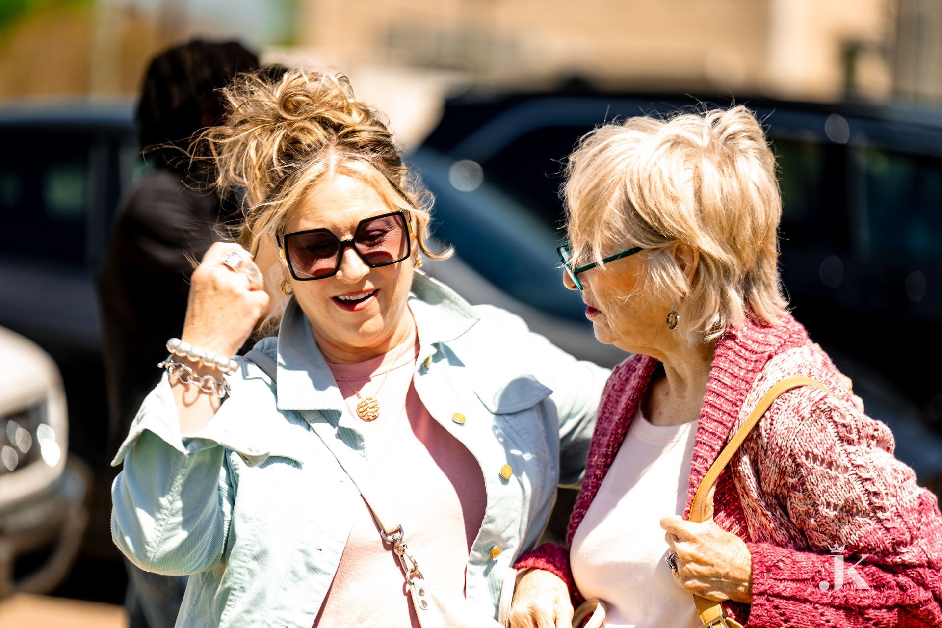 Two women are standing next to each other in a parking lot talking to each other.