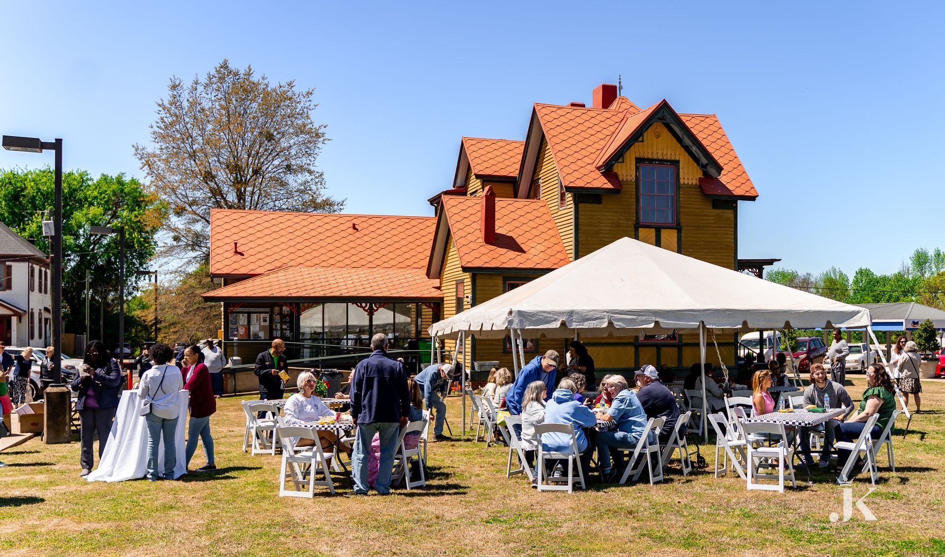 A group of people are sitting at tables under a tent in front of a large house.