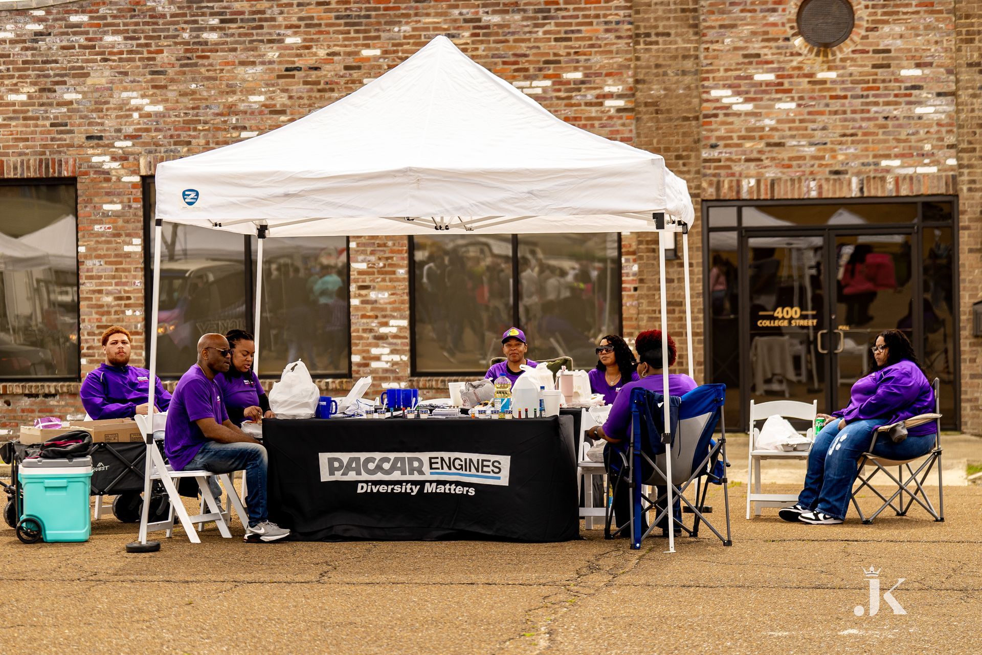 A group of people are sitting under a tent in front of a brick building.