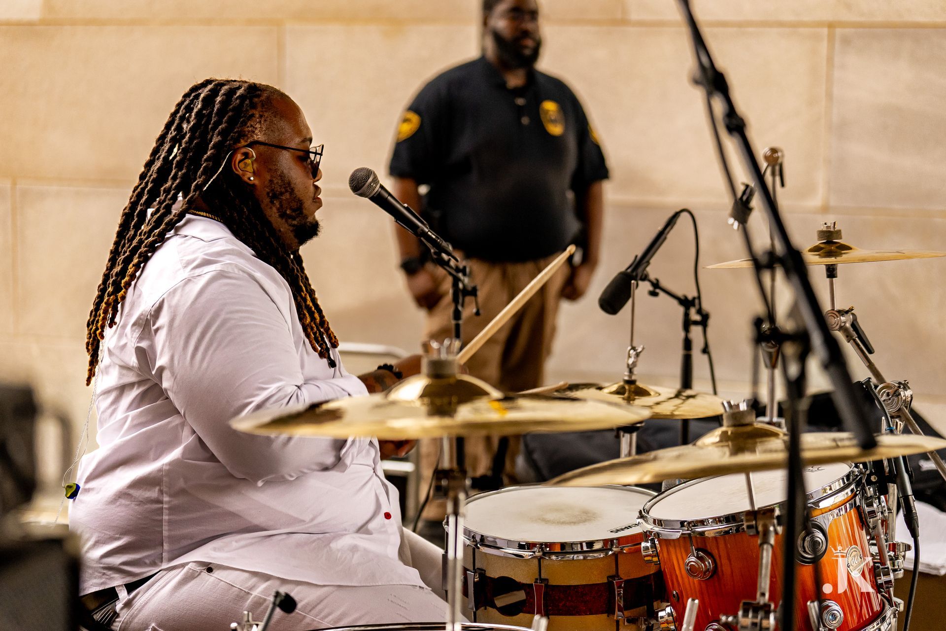 A man is playing drums in front of a microphone while another man stands behind him.