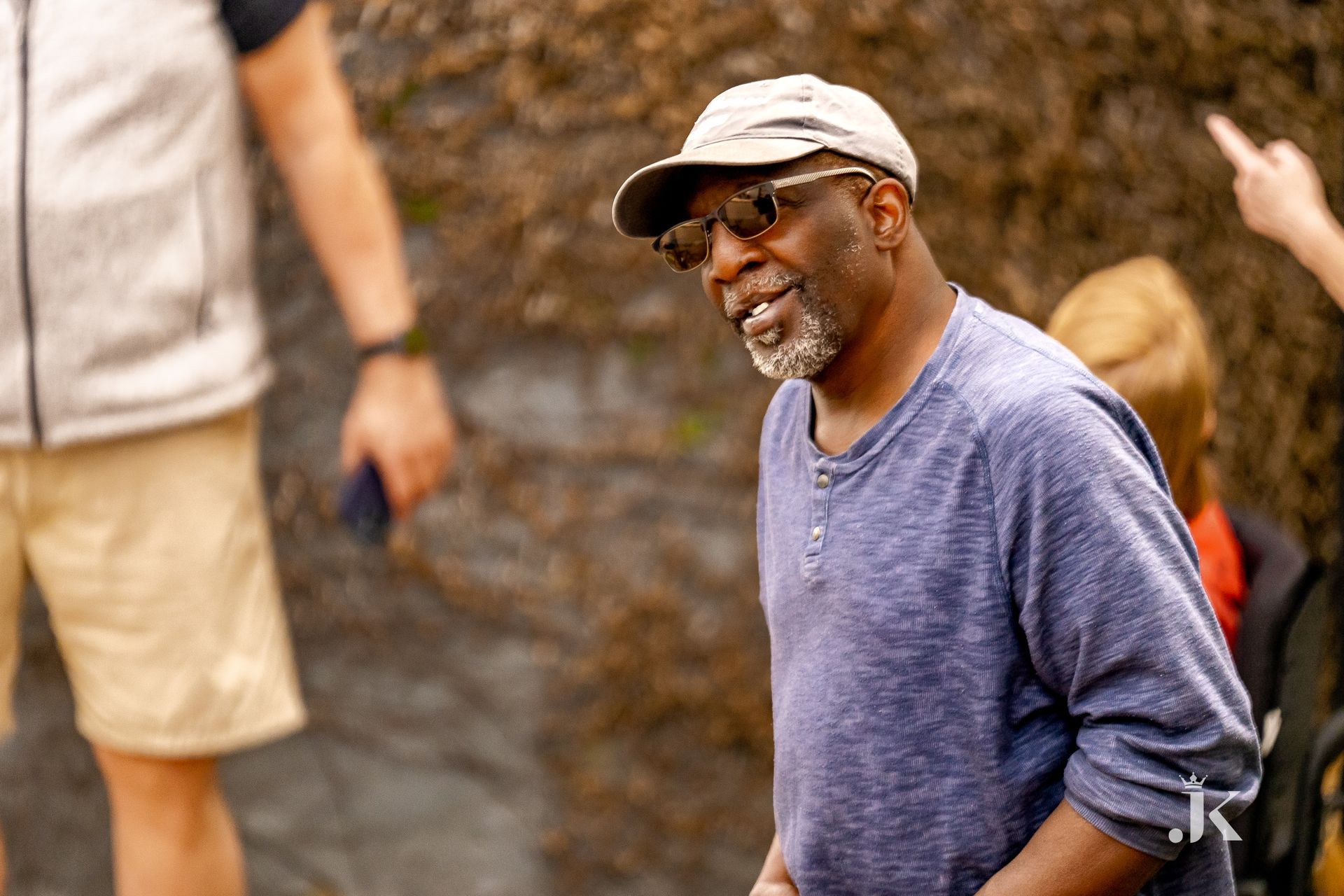 A man wearing a hat and sunglasses is standing in front of a group of people.