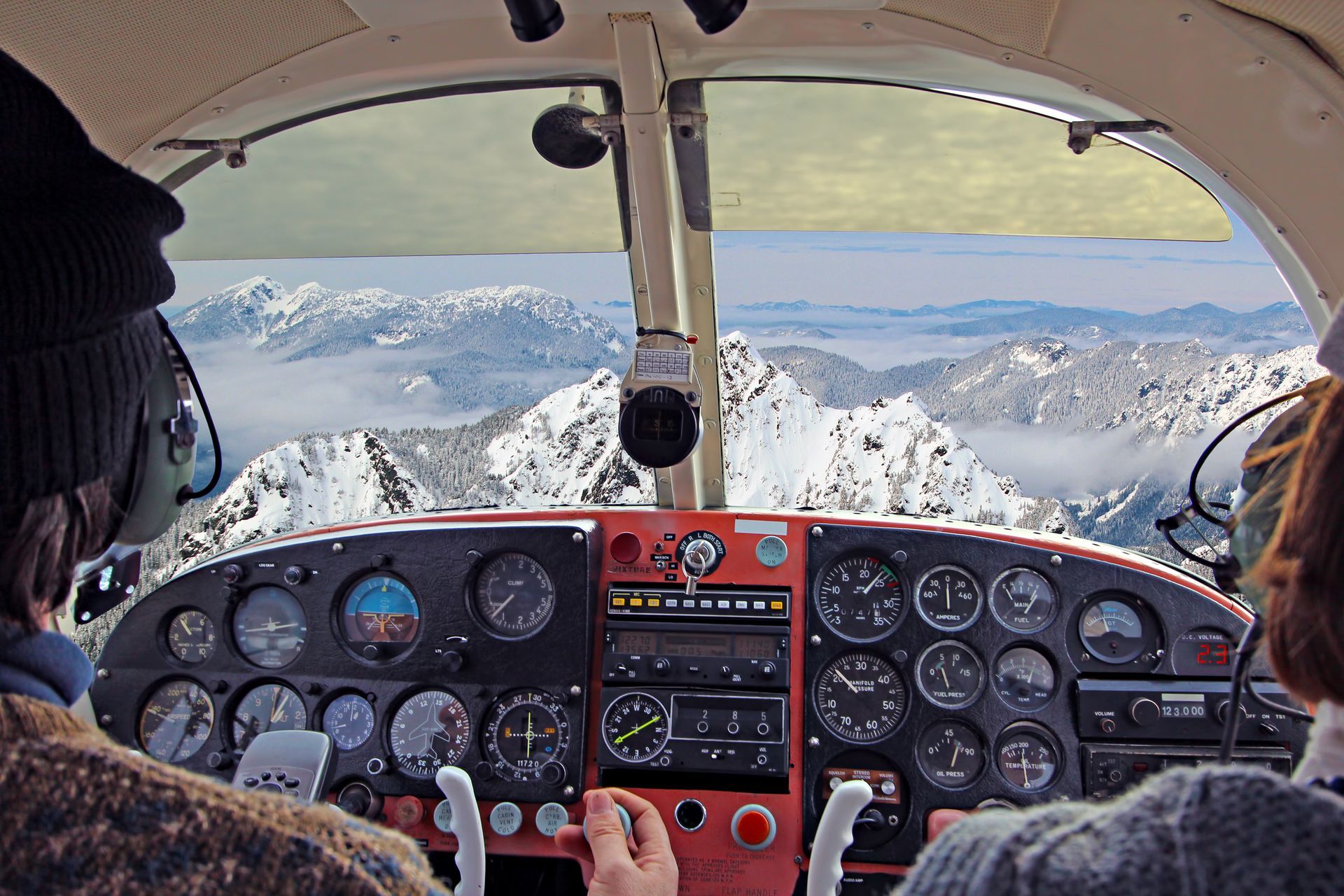 Two people are sitting in the cockpit of an airplane with mountains in the background
