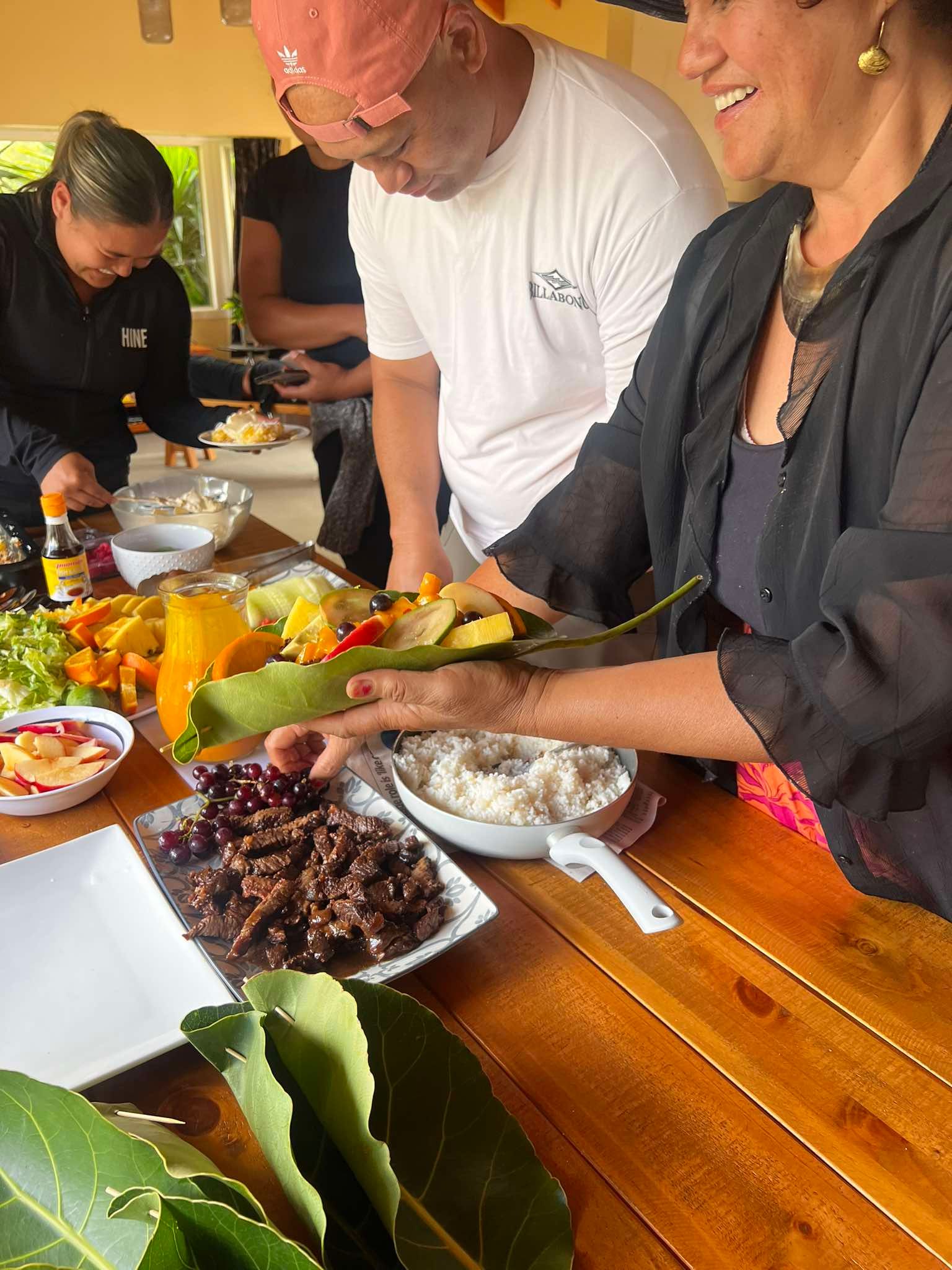 People preparing food on a wooden table outdoors. A woman smiles while holding a large leaf. Various dishes and ingredients are spread out.