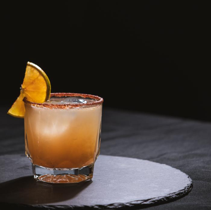 Cocktail in a glass with orange slice garnish on a dark stone surface, against a black background.