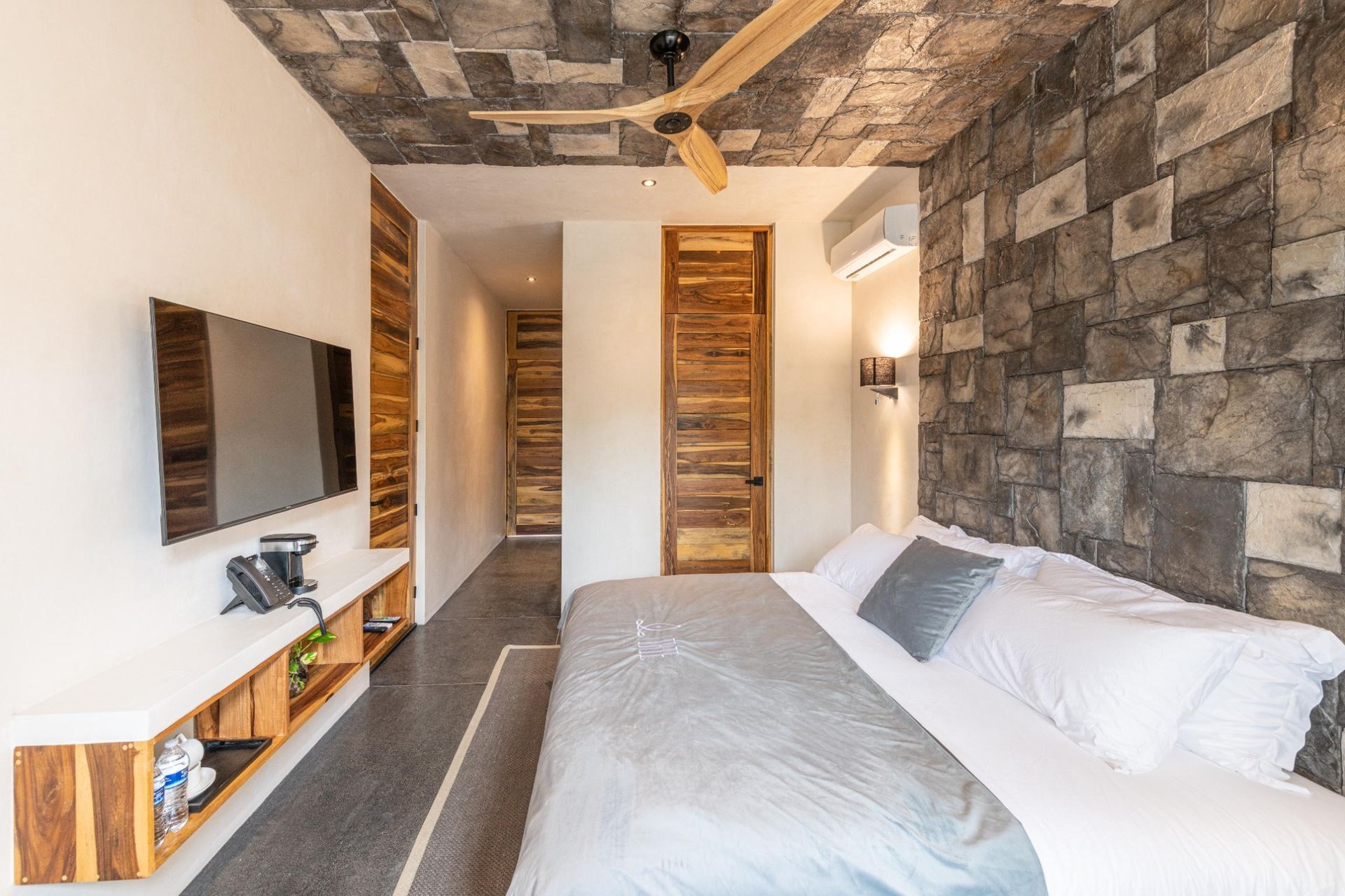 Bedroom with textured stone wall, wooden door and ceiling, white bed, TV, ceiling fan.