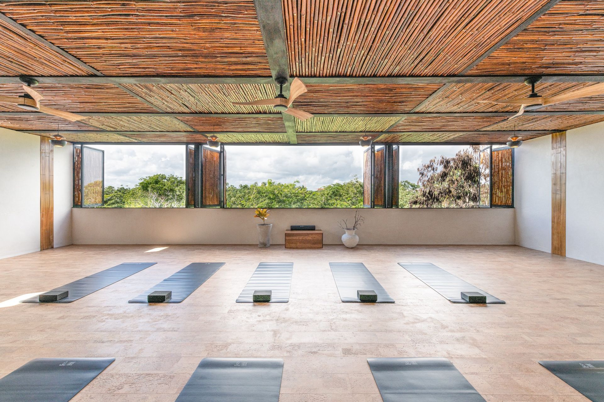 Yoga studio with mats, blocks, and open window overlooking greenery. Wooden ceiling and textured floor.