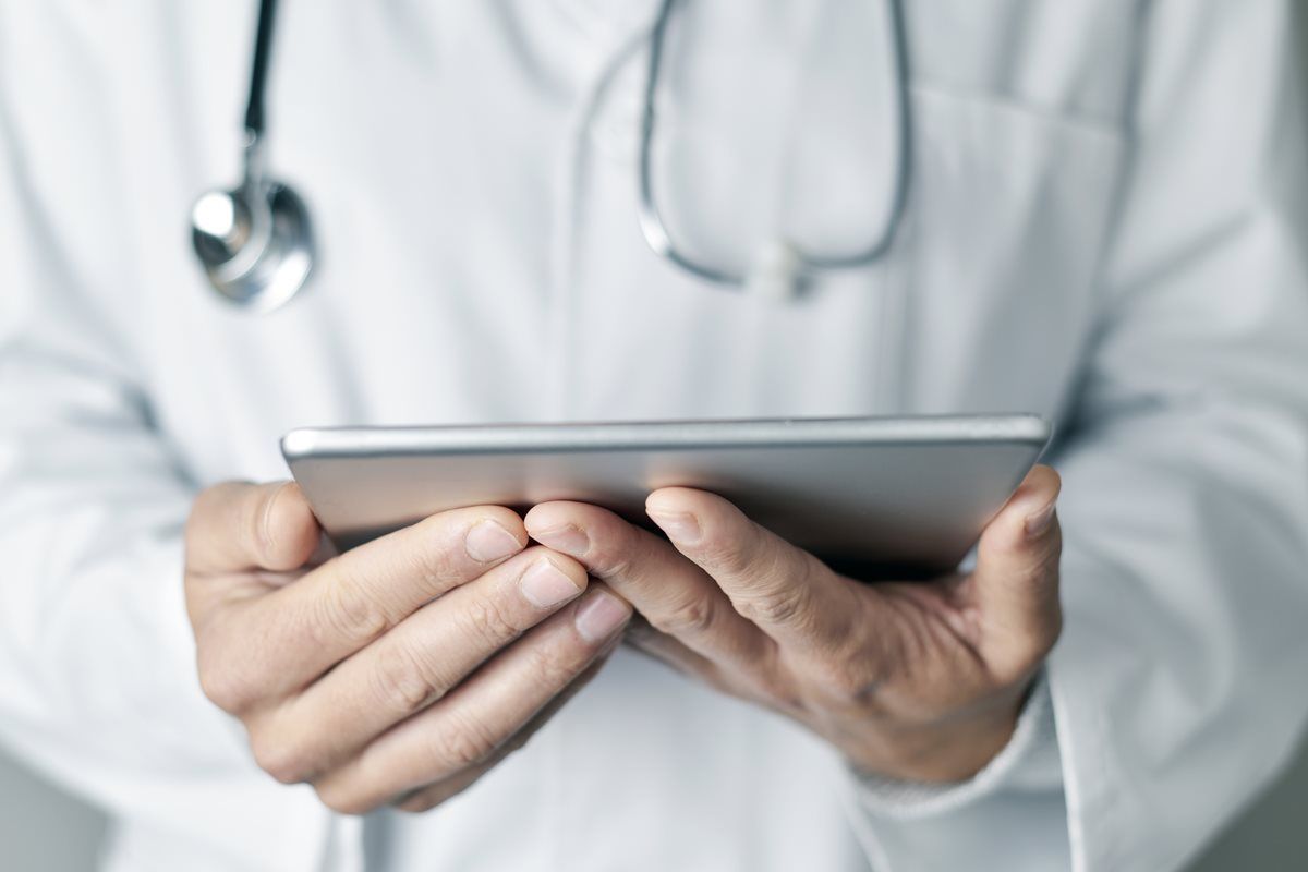A doctor with a stethoscope around his neck is holding a tablet in his hands.