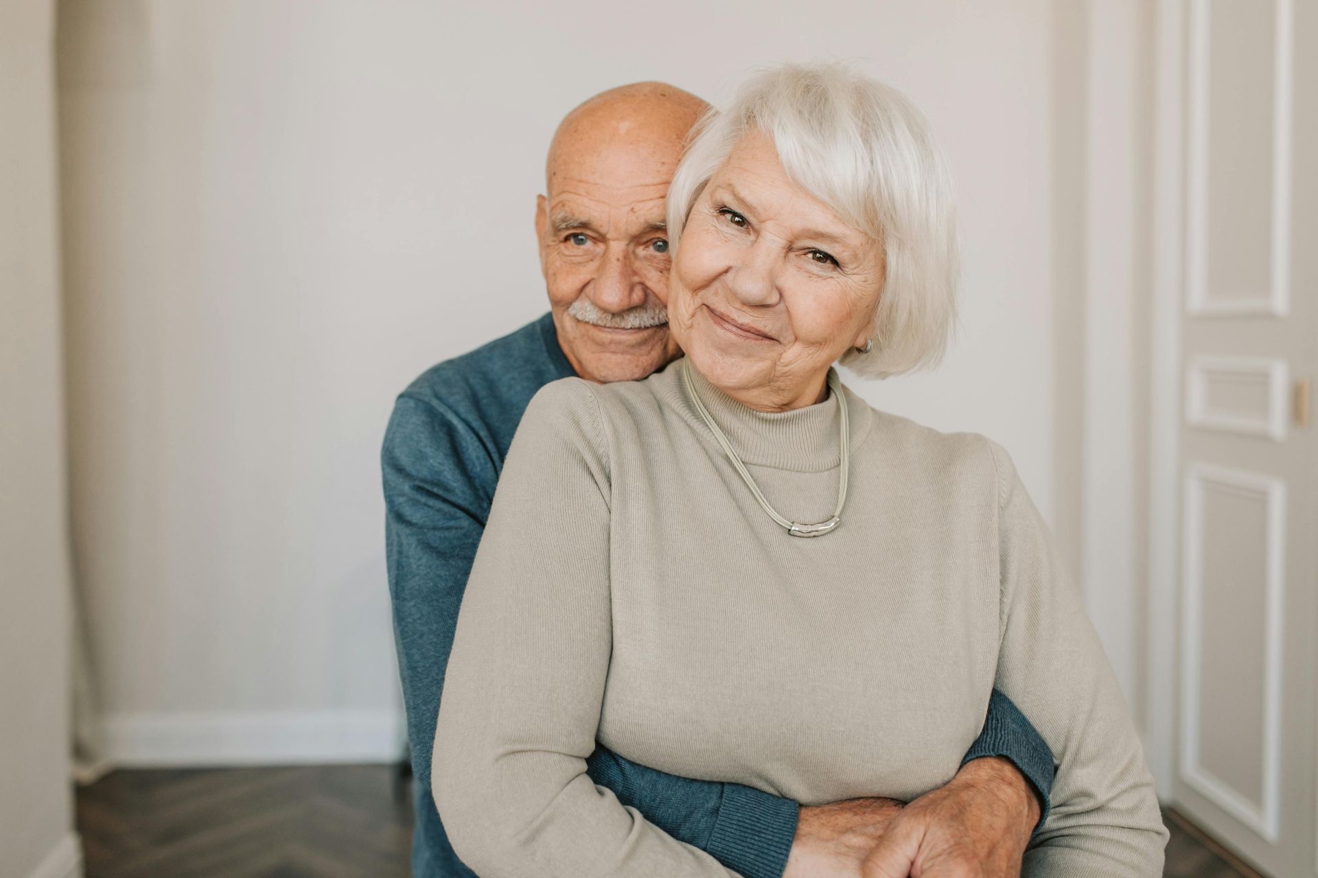 Senior couple embracing indoors, smiling. Man hugging woman from behind.