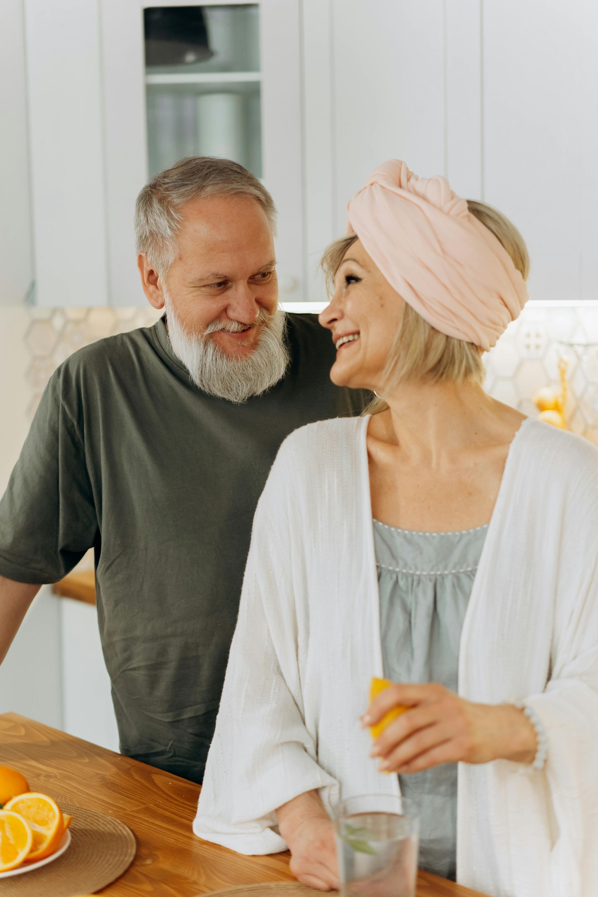 Elderly couple in kitchen, smiling. Woman in robe, headscarf; man with beard, green shirt. Citrus fruits on counter.