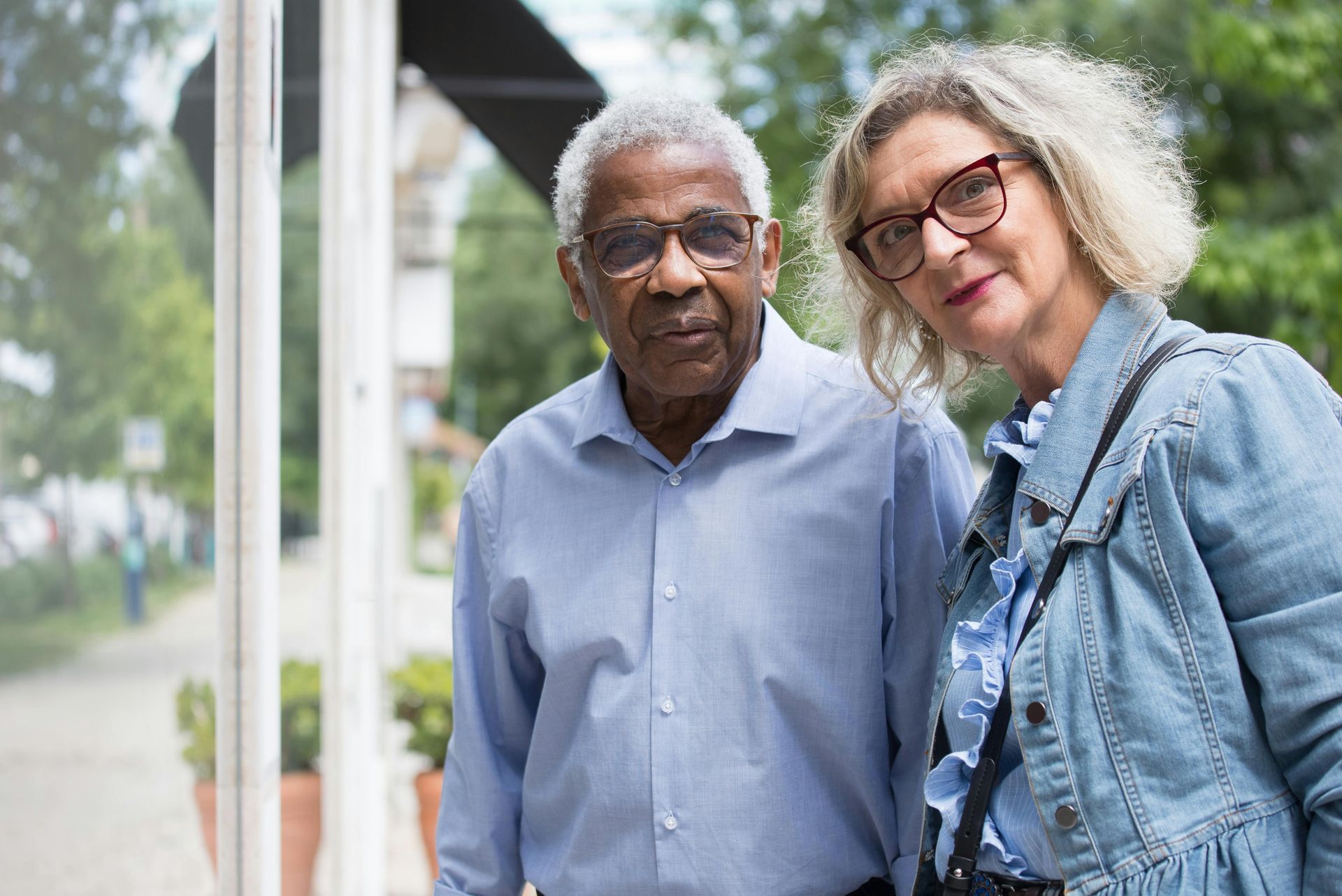 Two people, wearing glasses, stand outside. The man wears a blue shirt, the woman a denim jacket.